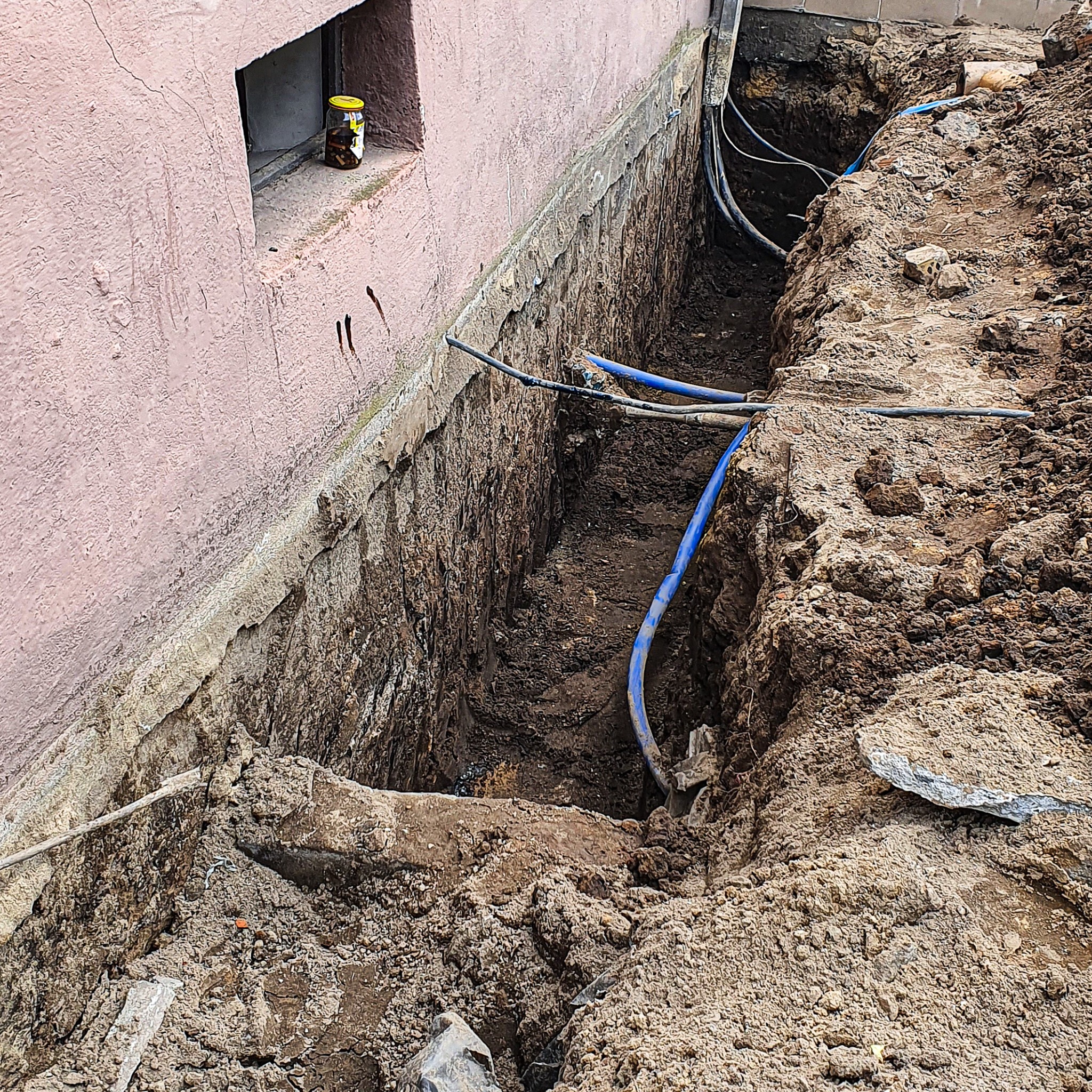 Wykop wzdłuż fundamentu budynku z widocznymi kablami i rurami, słoik z zawartością w okienku piwnicznym.