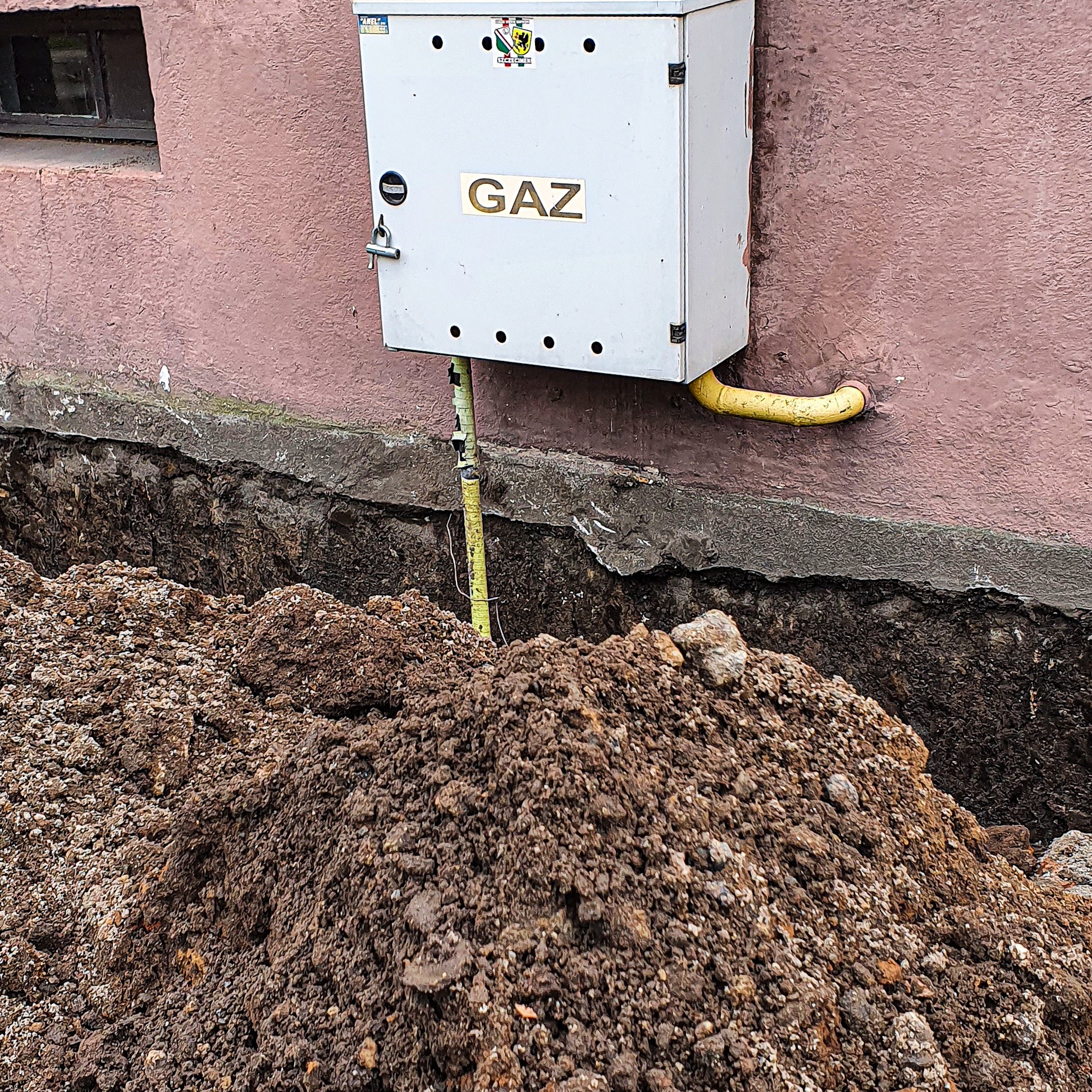 Wykonany wykop w ziemi przy ścianie budynku z instalacją gazową, widoczna skrzynka z napisem GAZ i żółta rura gazowa.