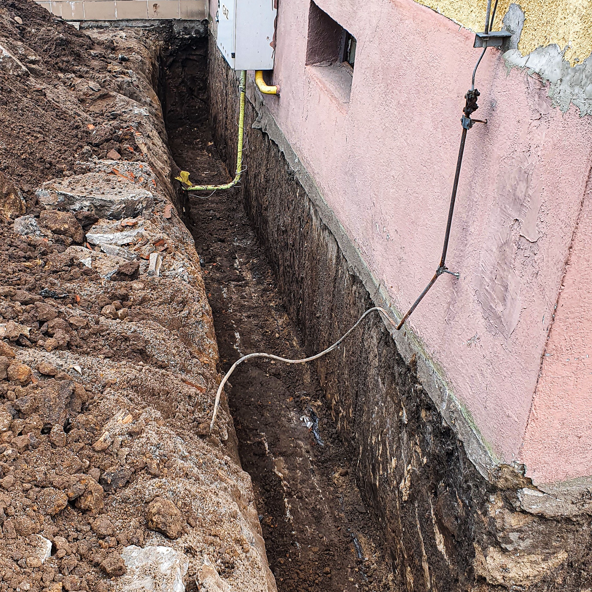 Wykopany wąski rów wzdłuż fundamentu budynku z widocznymi instalacjami: rura gazowa i kabel elektryczny, ziemia usypana po jednej stronie.