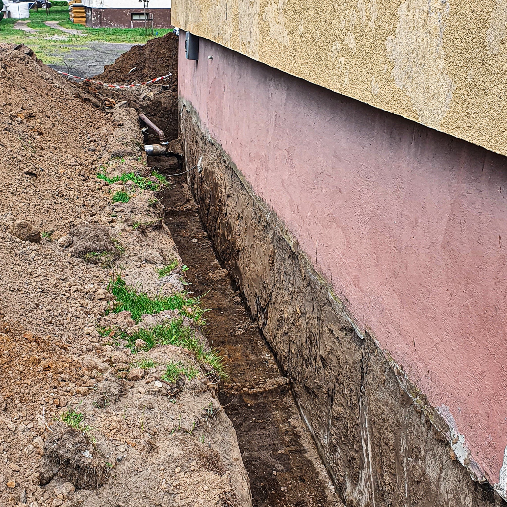 Wąski wykop wzdłuż fundamentu budynku z widocznymi rurami, zabezpieczony taśmą ostrzegawczą, obok pryzma ziemi.