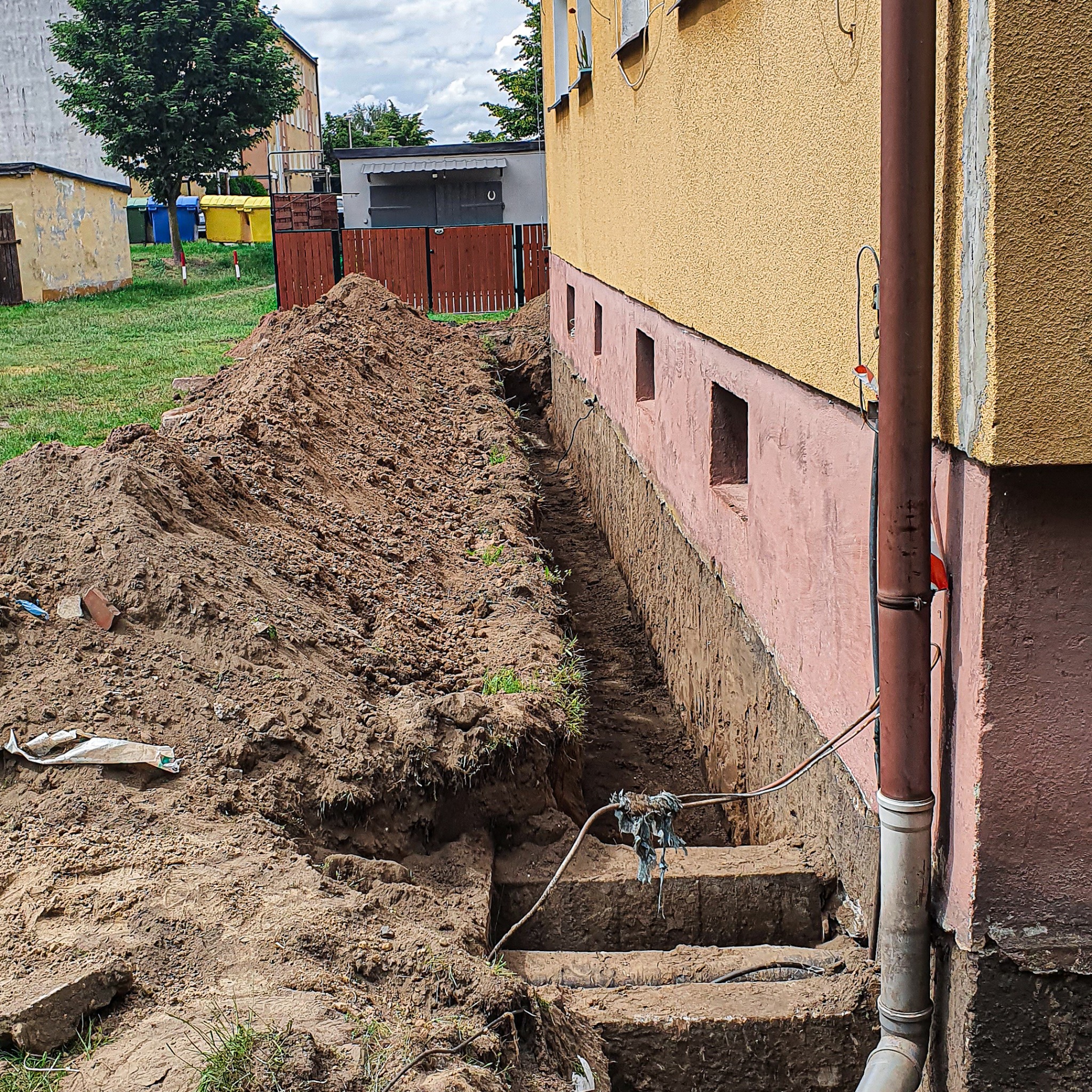 Wykop wzdłuż fundamentu budynku z widoczną ziemią, schodkowym zejściem i wystającymi przewodami.