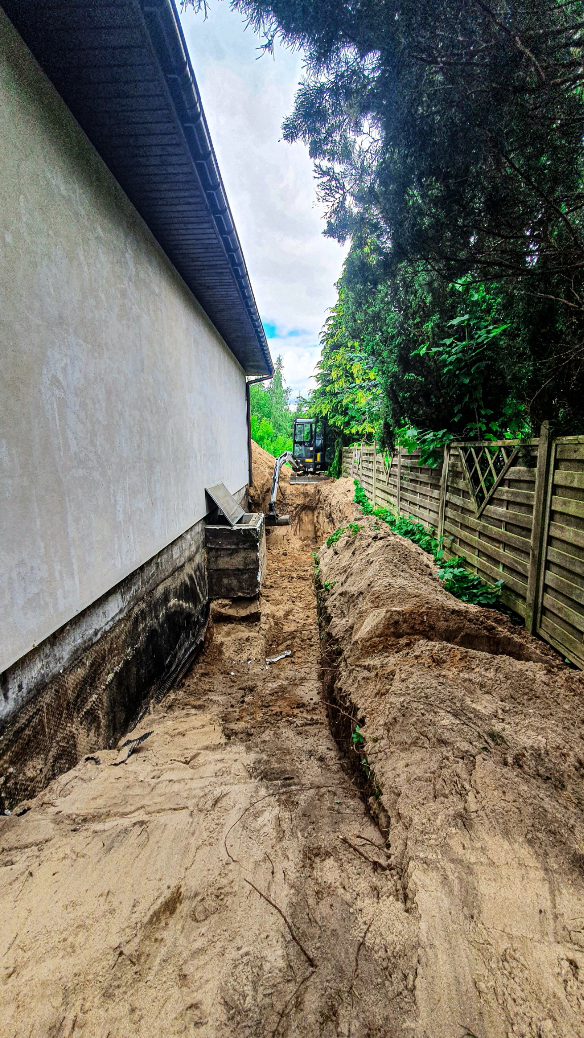 Wąski wykop w piasku wzdłuż ściany budynku, wykonany przy użyciu małej koparki widocznej w tle, ogrodzenie drewniane po prawej stronie.
