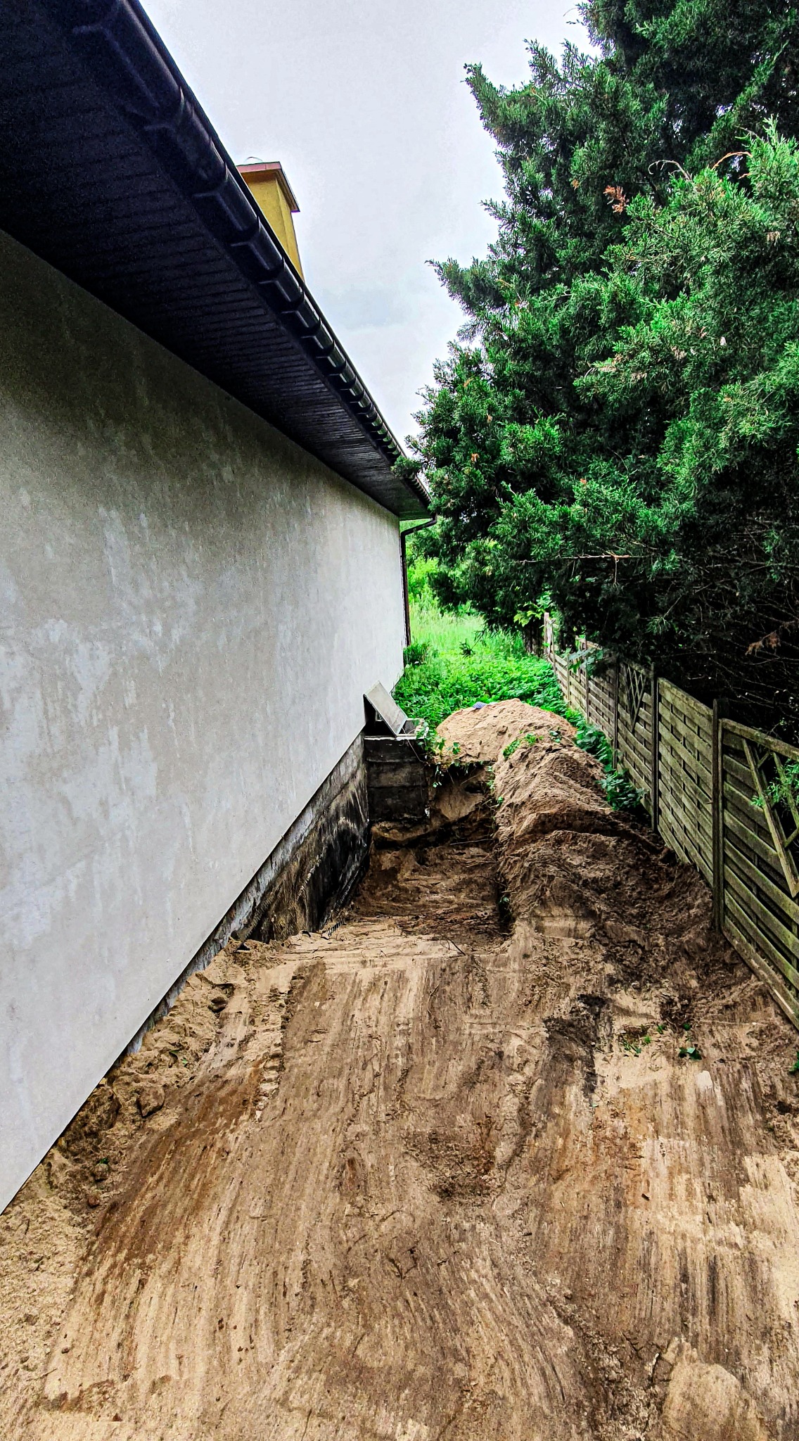 Wykop wzdłuż ściany budynku z widocznymi śladami po pracy koparki, częściowo zasypany ziemią, z drewnianym ogrodzeniem w tle.