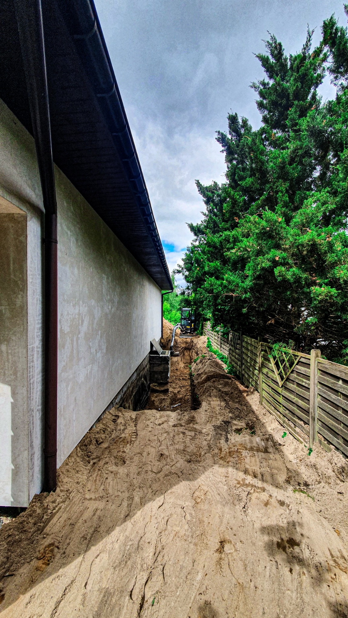 Wąski wykop wzdłuż ściany budynku, zabezpieczony szalunkiem, z widocznym minikoparką w oddali, ziemia usypana wzdłuż wykopu, obok drewniany płot i wysokie drzewo.