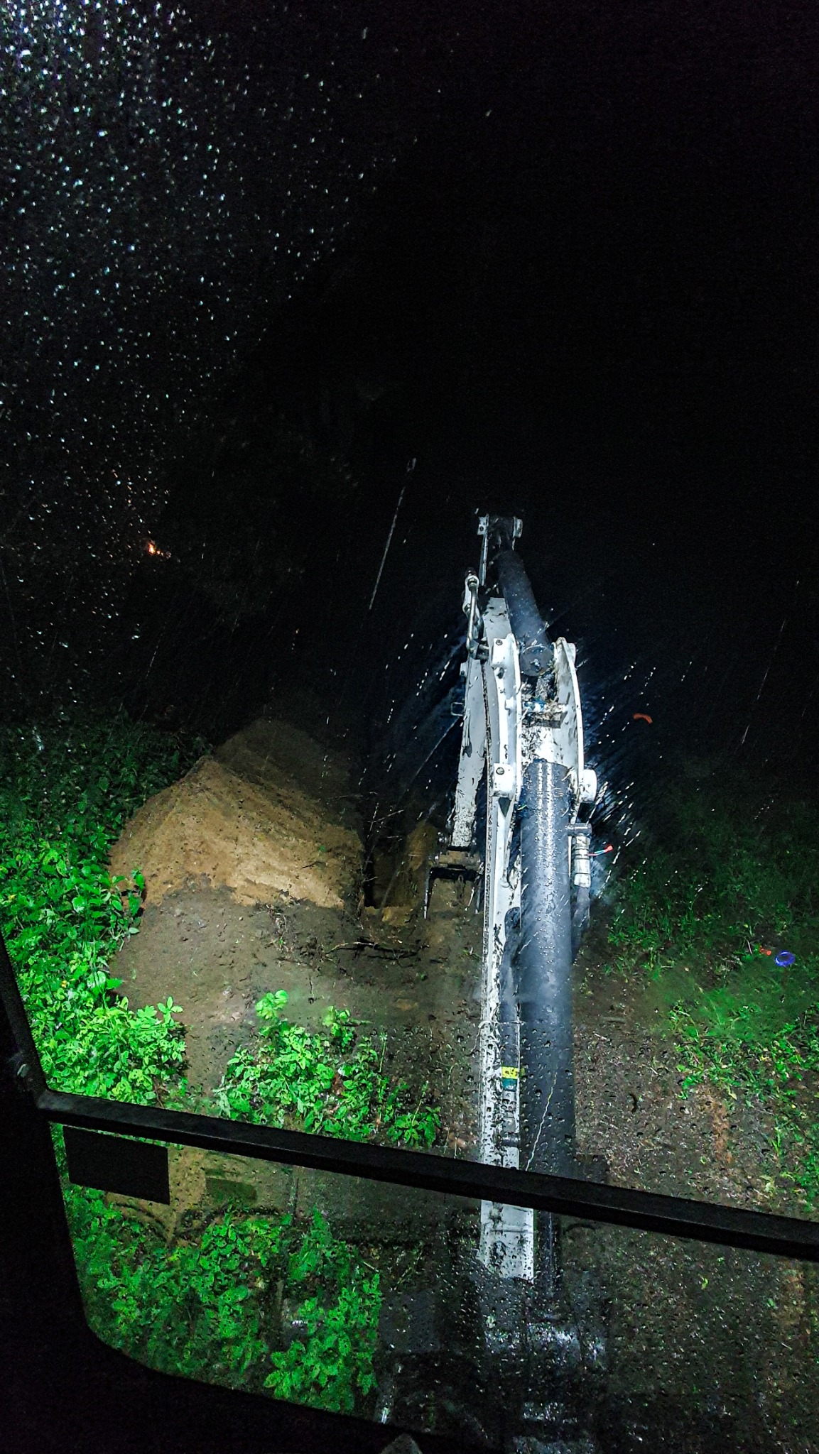 Widok z kabiny na białą koparkę pracującą w nocy w deszczu, widoczny wykop w ziemi i bujna roślinność.