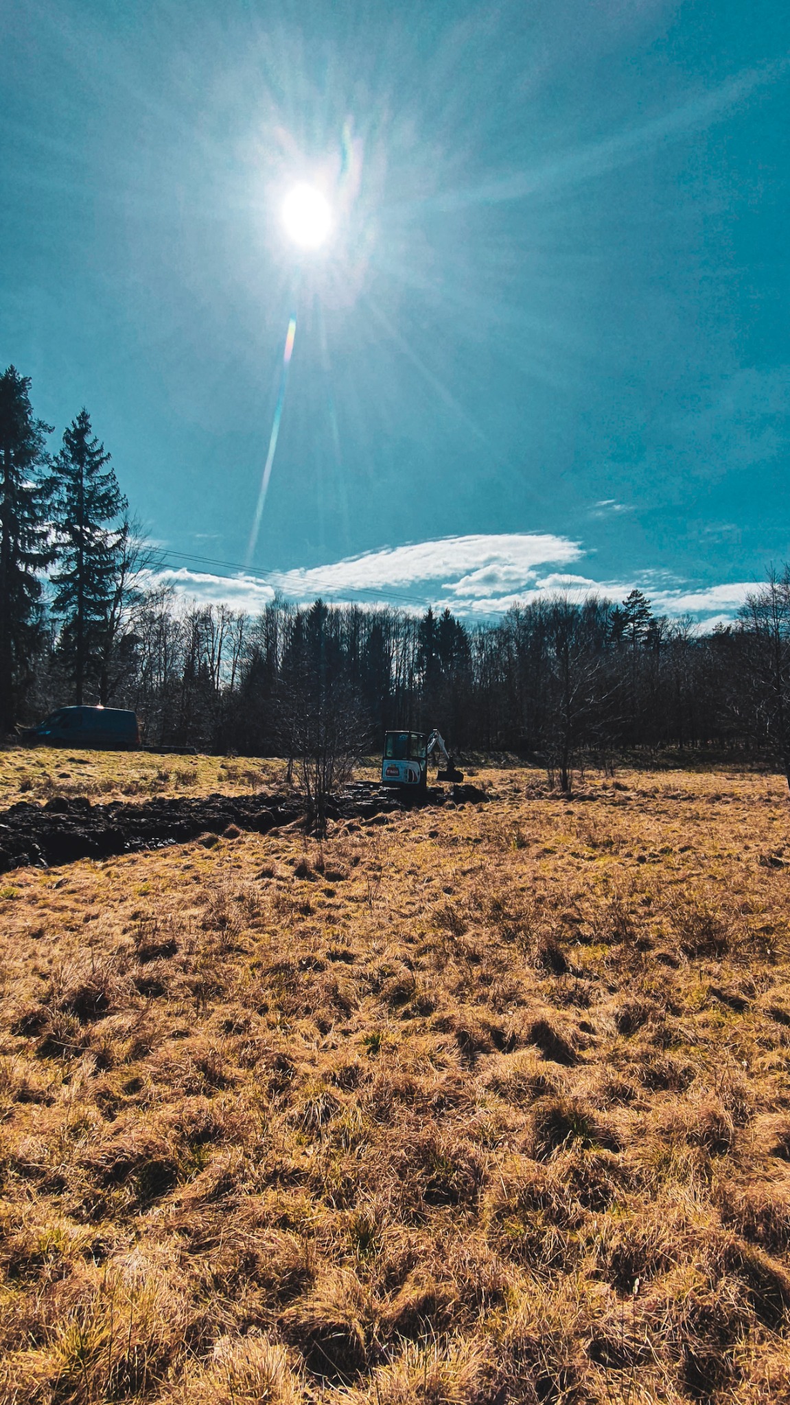 Mała koparka pracuje na zewnątrz, w tle las i błękitne niebo z silnym słońcem, świeżo wykopany rów w trawiastym terenie.