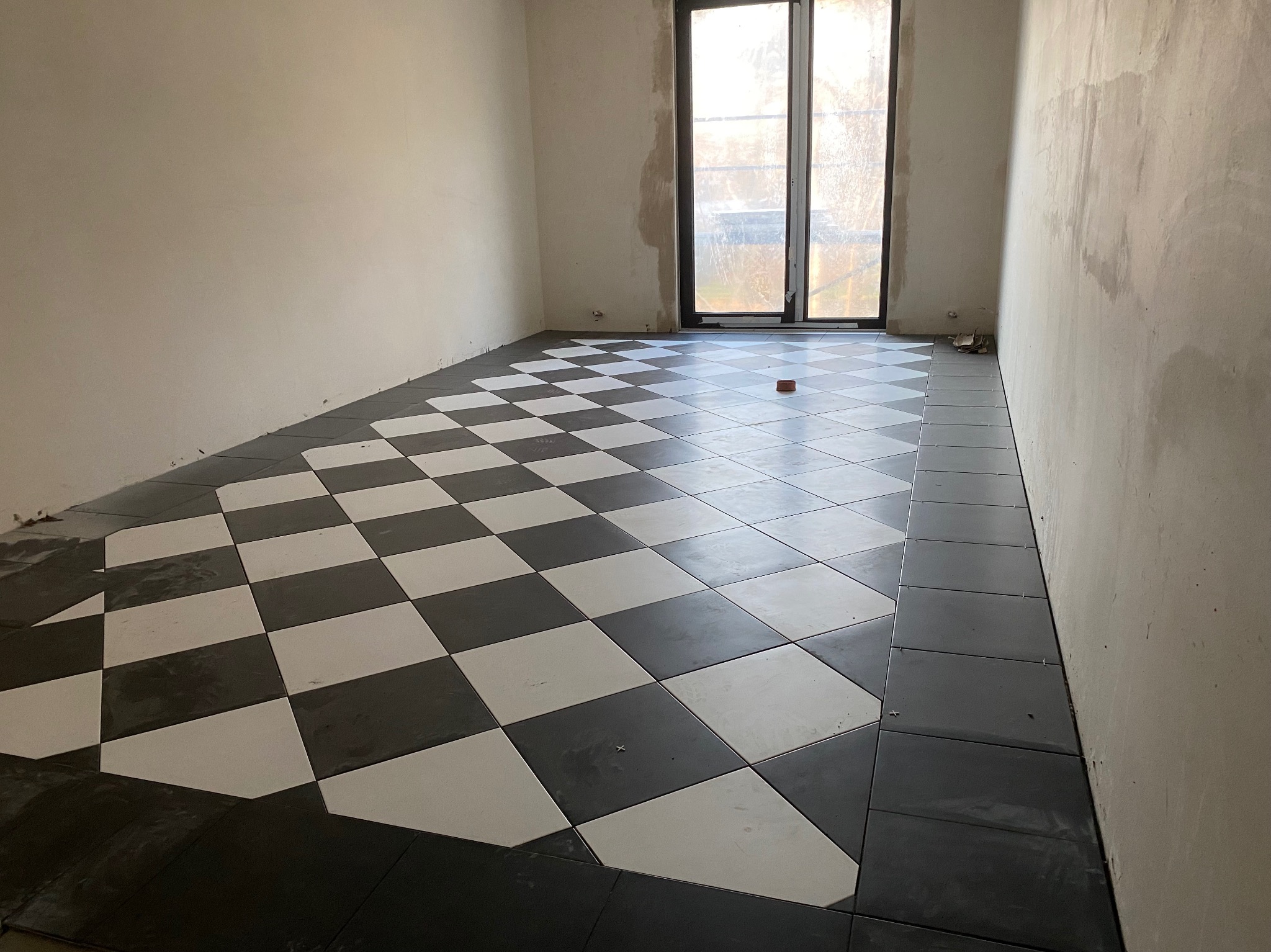 Układanie czarno-białej szachownicy z płytek na podłodze w pokoju, widoczne niedokończone krawędzie i okno w tle.