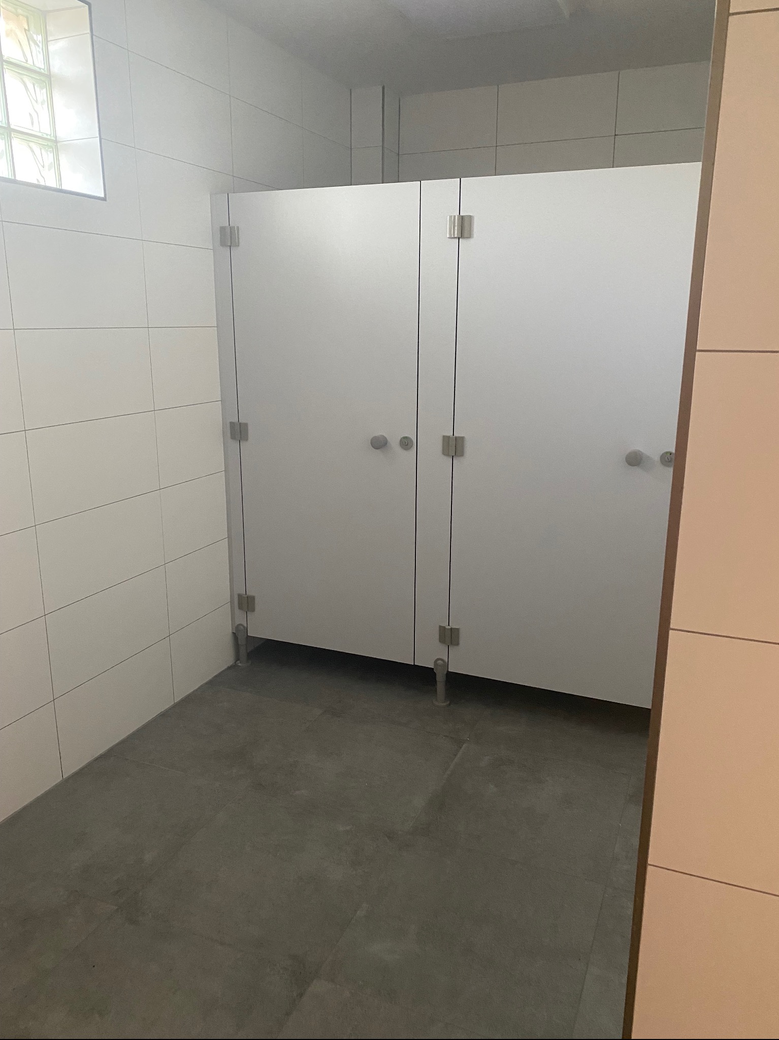 Wnętrze publicznej toalety z kabinami wykończonymi białymi płytkami i szarymi płytkami podłogowymi, widoczne metalowe okucia kabin i szklany luksfer w oknie.