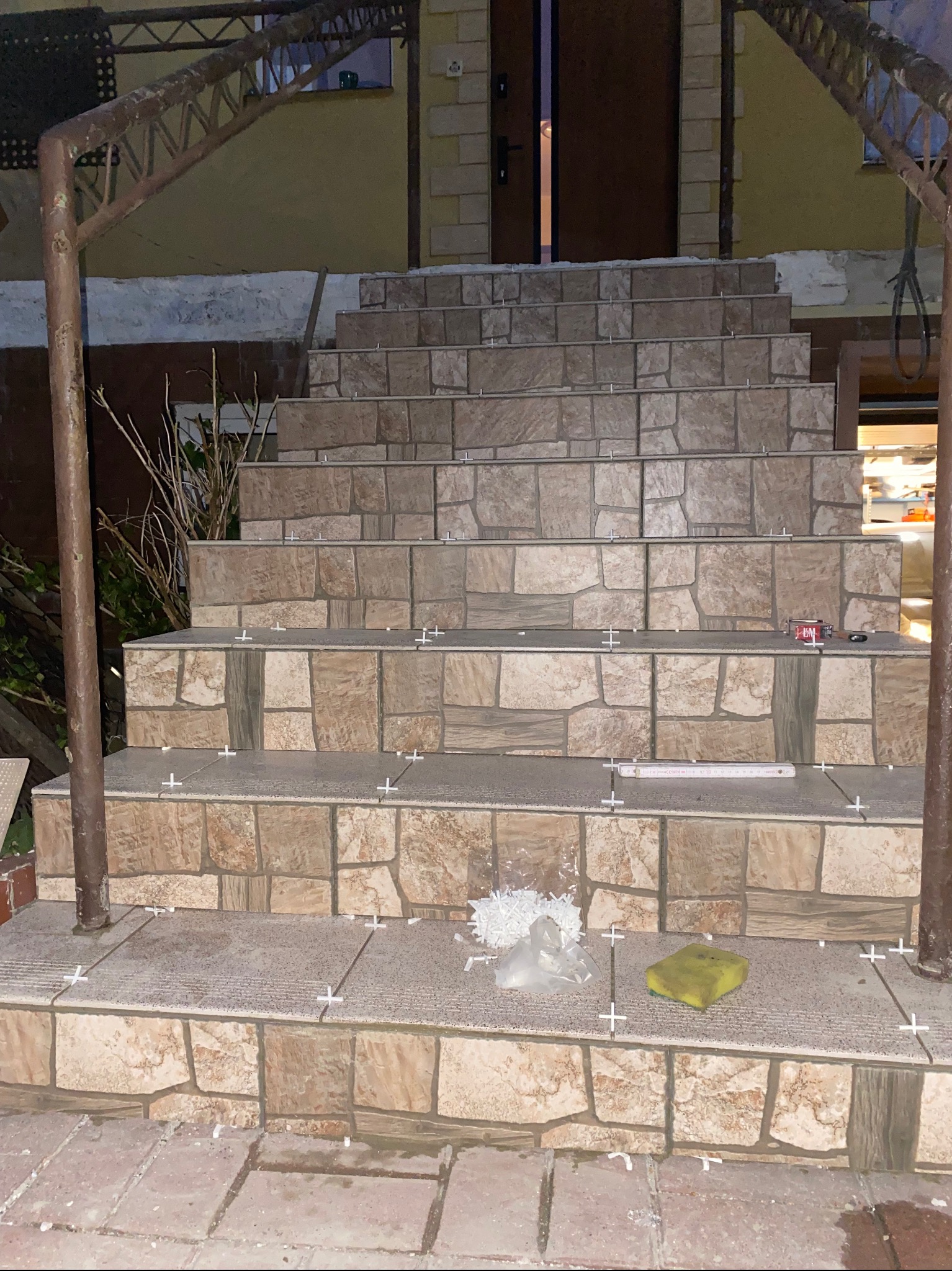 Wyłożone płytkami schody zewnętrzne w trakcie prac, widoczne krzyżyki dystansowe, gąbka i opakowanie po fugach.