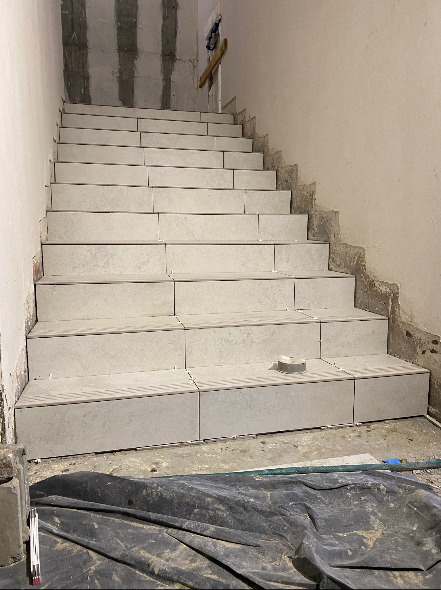 Schody w trakcie układania płytek ceramicznych, widoczne szare płytki na stopniach, plastikowe krzyżyki dystansowe, taśma malarska, folia ochronna na podłodze i surowe ściany w tle.