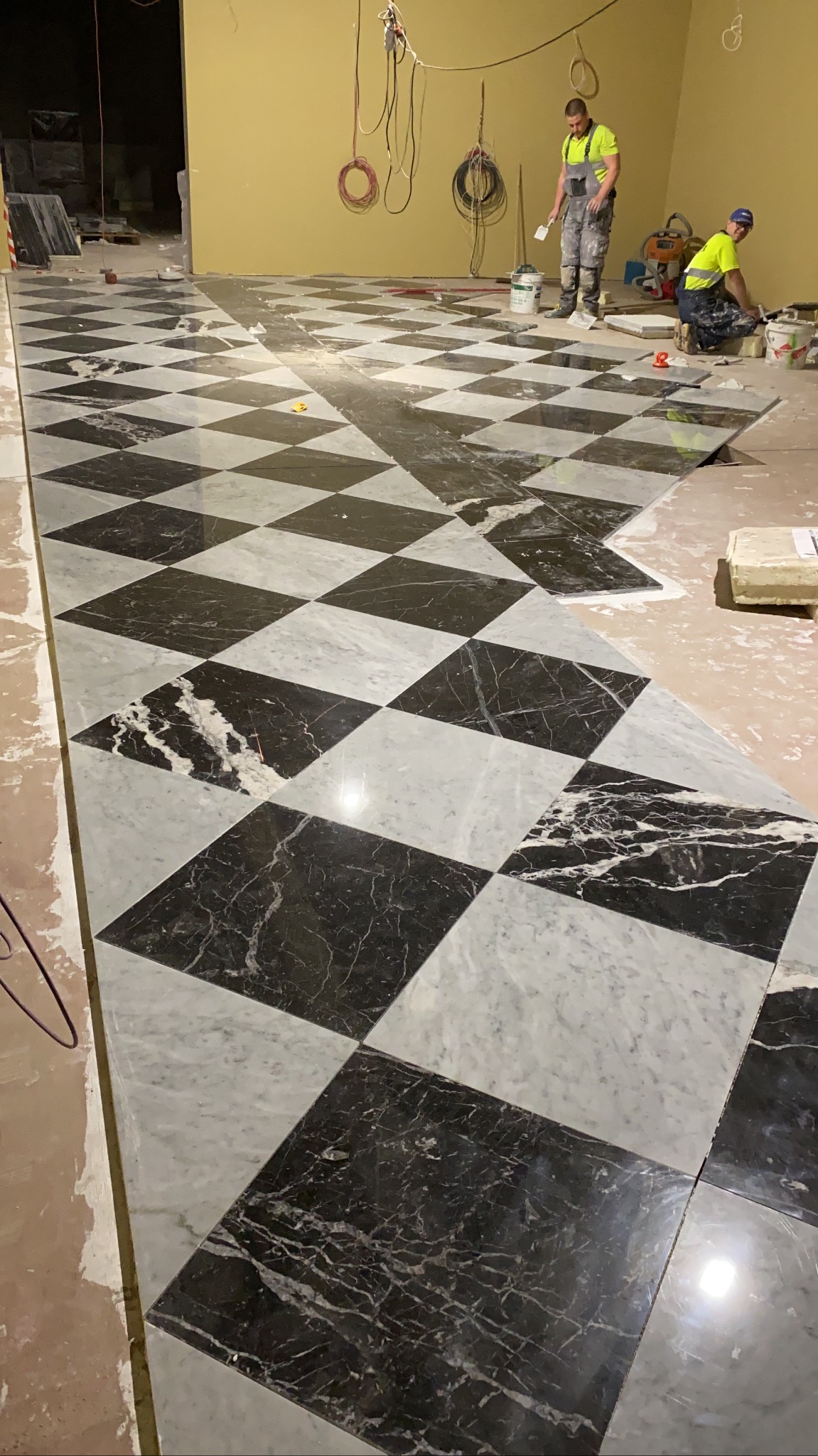 Układanie podłogi z czarno-białych marmurowych płytek w szachownicę; dwóch pracowników w strojach roboczych w tle, jeden stojący z pacą, drugi klęczący przy wiadrze z zaprawą.