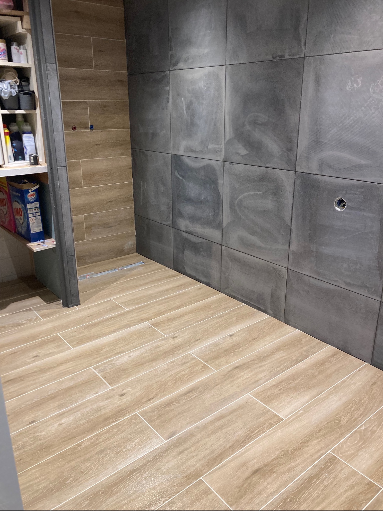 Wyłożona kafelkami łazienka z szarymi płytkami na ścianach i jasnymi, drewnopodobnymi płytkami na podłodze, widoczna półka z detergentami w tle.