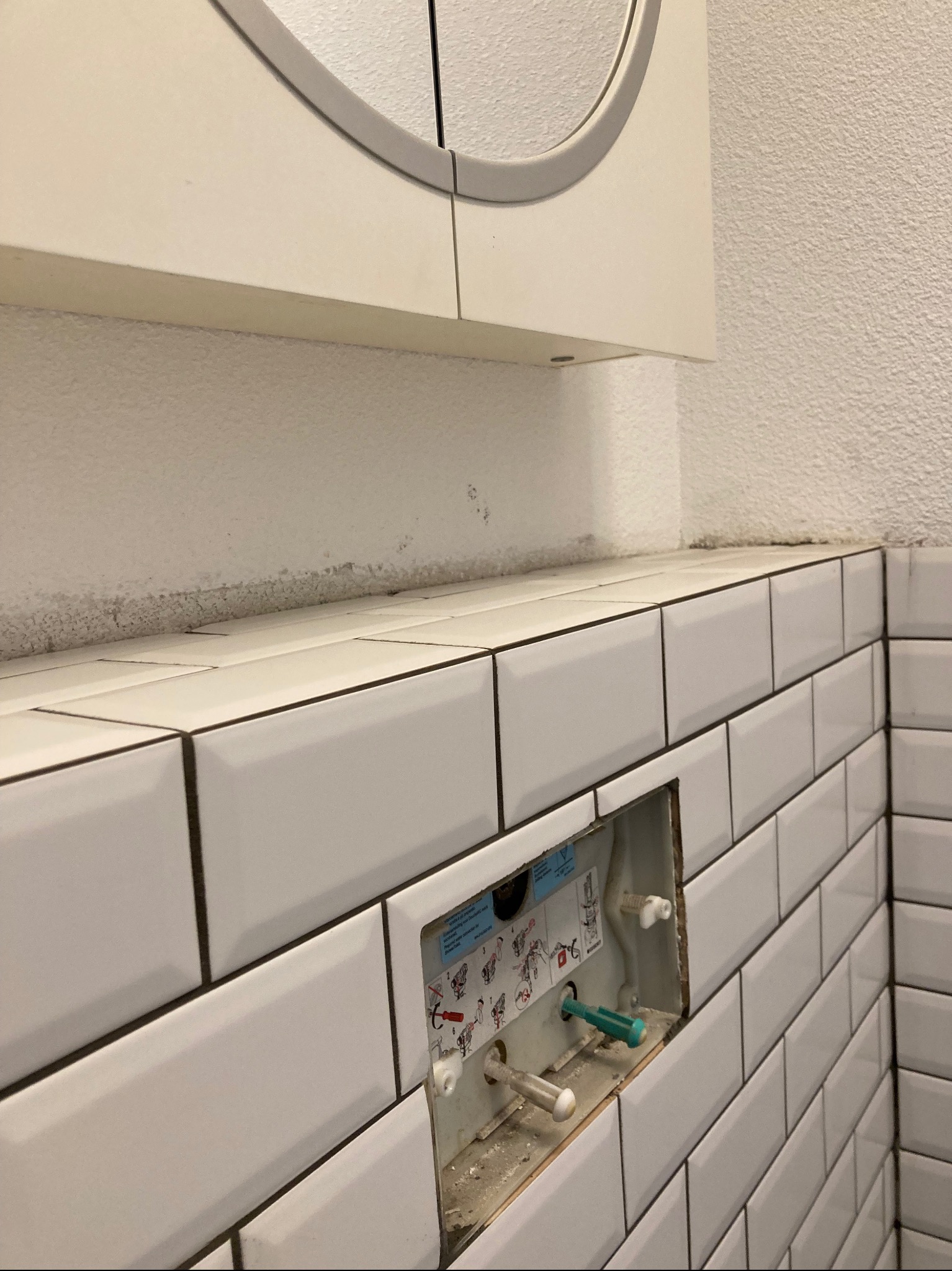 Białe, prostokątne płytki ułożone na ścianie, z widoczną instalacją podtynkową do toalety w trakcie montażu. Widoczne elementy mocujące i instrukcja montażu na obudowie.