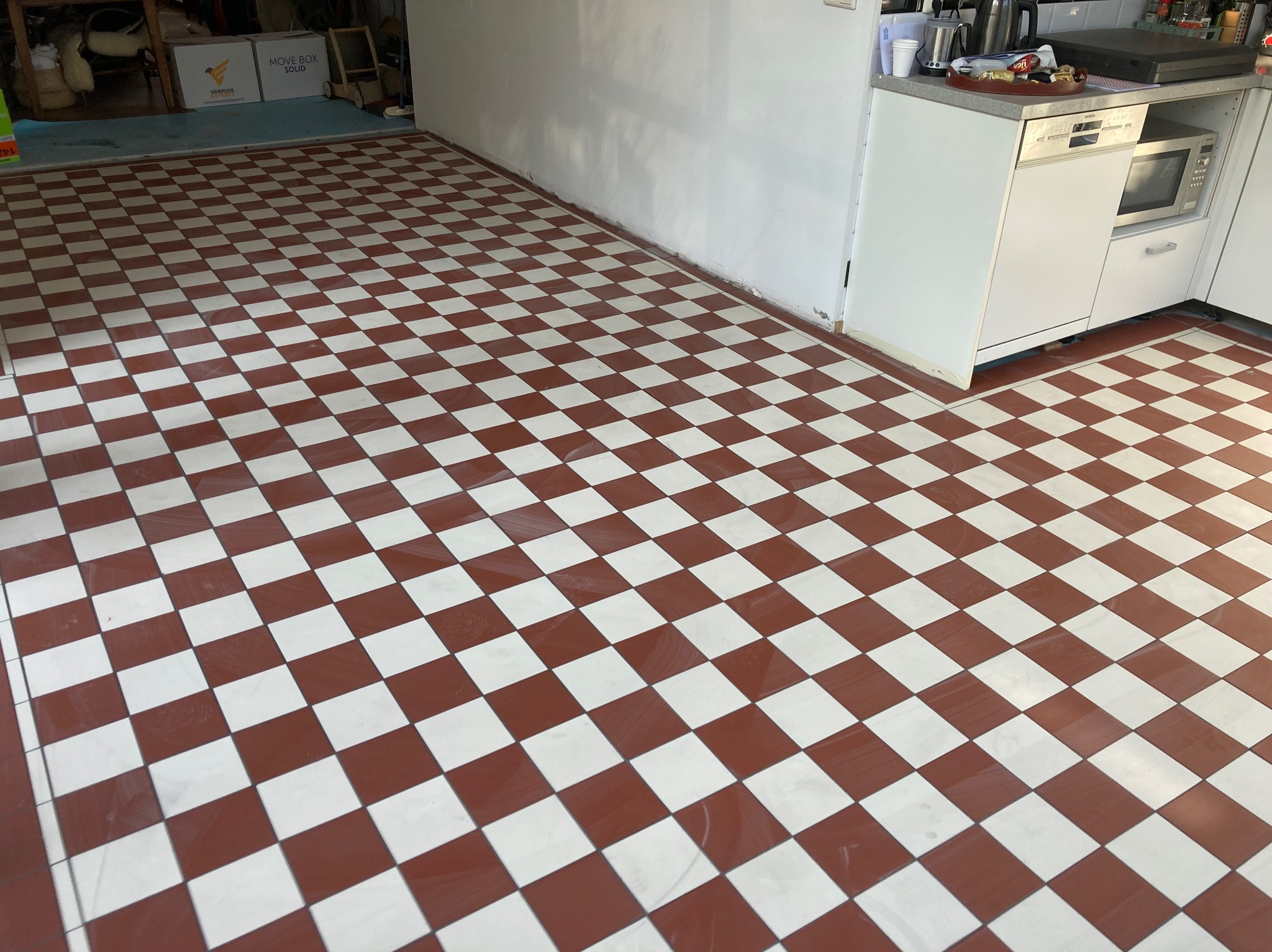 Czerwono-biała podłoga w szachownicę z płytek ceramicznych, widoczna w kuchni przy białych szafkach, z lekkim odblaskiem światła na powierzchni.