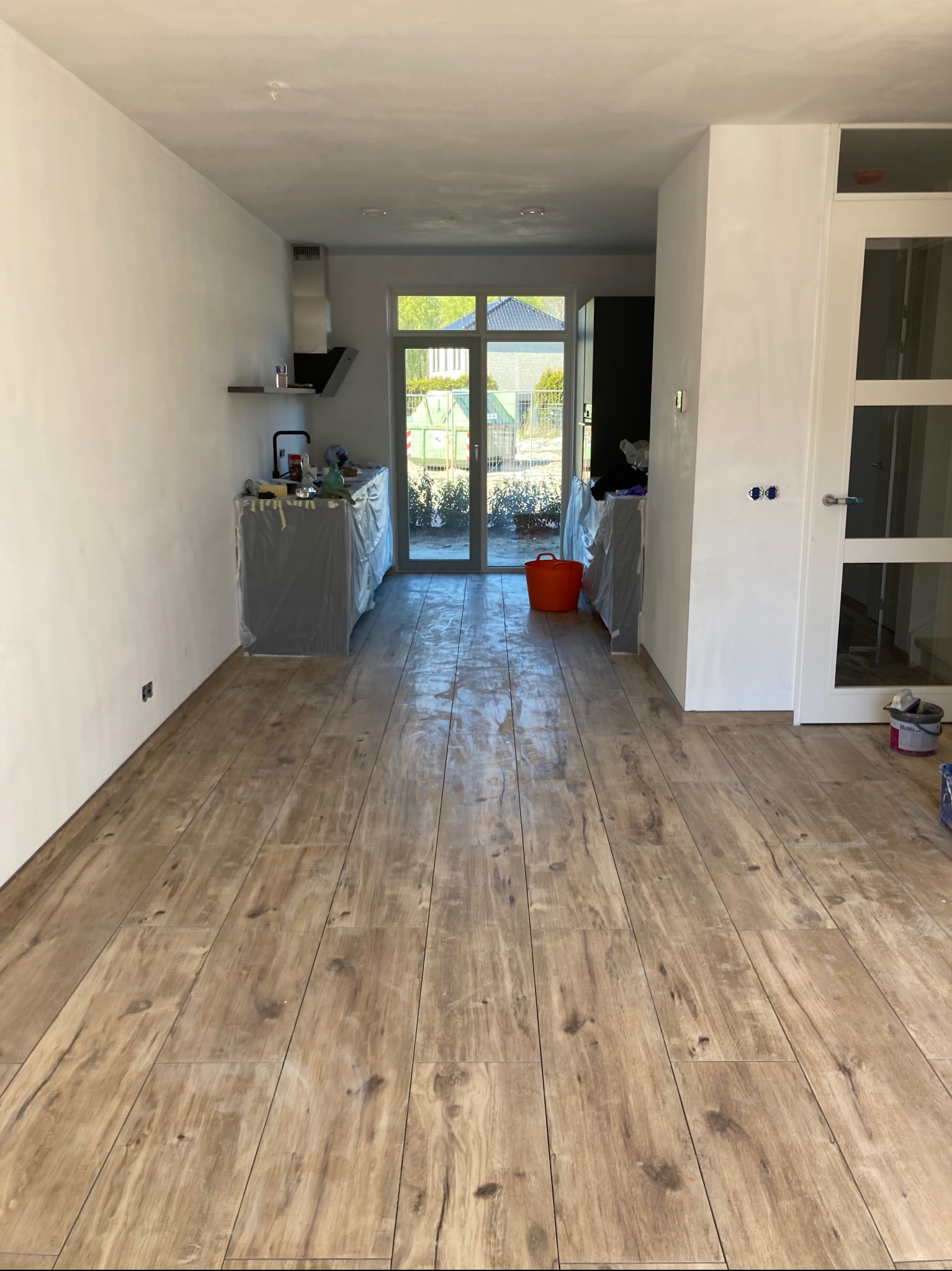 Ułożona podłoga z płytek imitujących drewno w nowym, jasnym wnętrzu z widocznymi zabezpieczonymi meblami i otwartym przejściem do ogrodu.