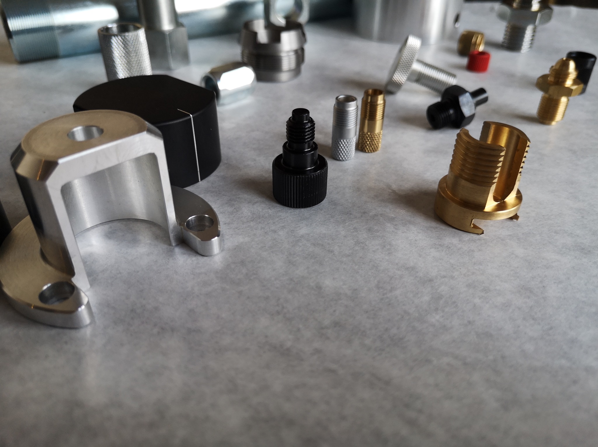Różnorodne, precyzyjnie wykonane metalowe elementy o różnych kształtach i wykończeniach, ułożone na szarym tle. Widoczne detale gwintów, frezów i otworów. Części wykonane z aluminium, mosiądzu...