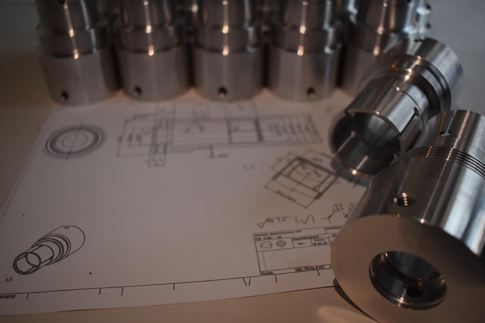 Srebrne, metalowe elementy cylindryczne z gwintem leżące na planie technicznym, widoczne detale obróbki skrawaniem i otwory.