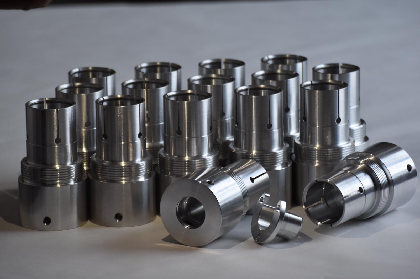 Grupowe ujęcie precyzyjnie obrobionych, metalowych tulejek z rowkami i otworami, leżących na jasnym tle, ukazujące detale frezowania i gwintowania.