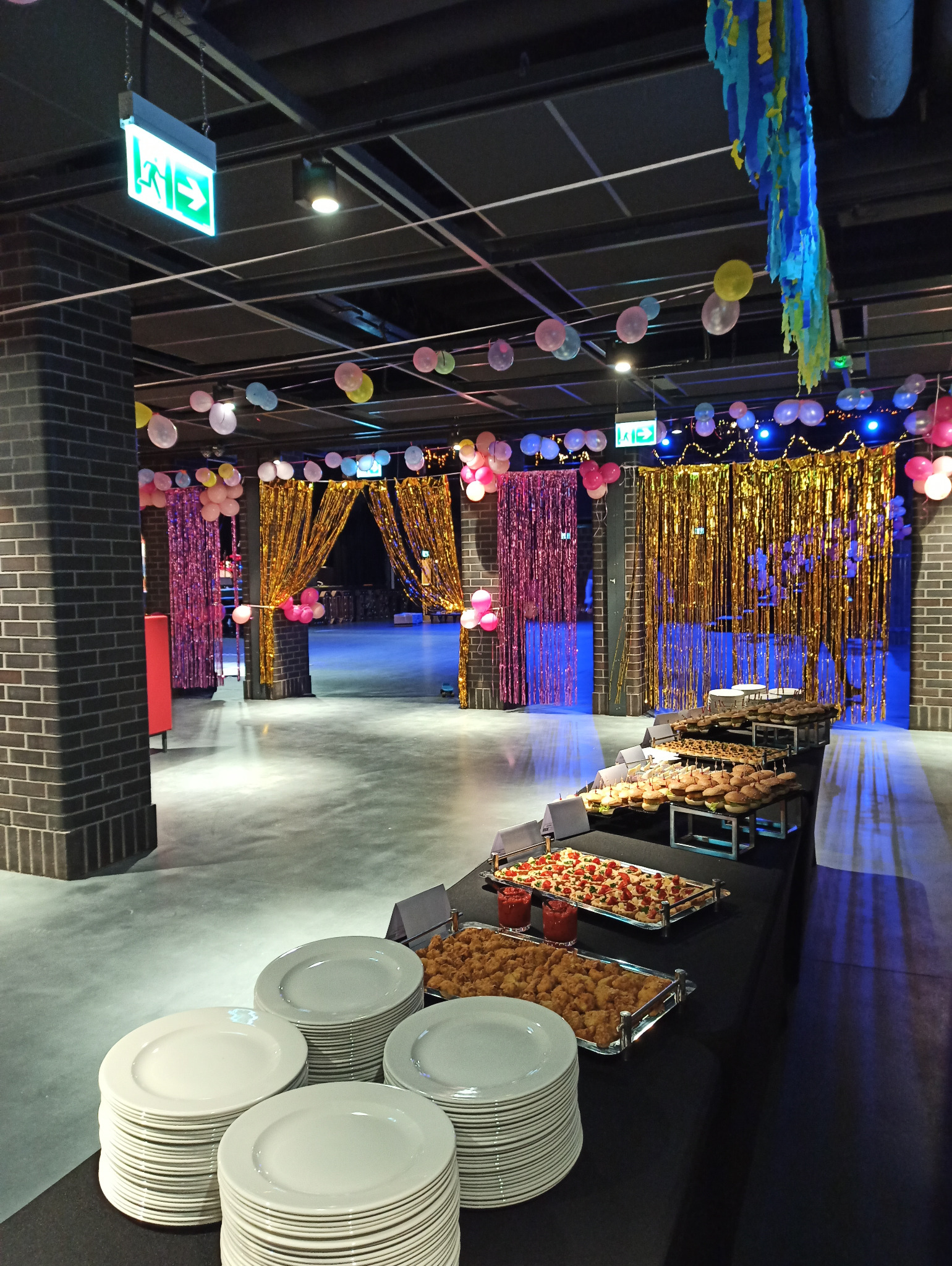 Uroczyste wnętrze z dekoracjami balonowymi i złotymi kurtynami, zastawiony stół z przekąskami i stosami białych talerzy.