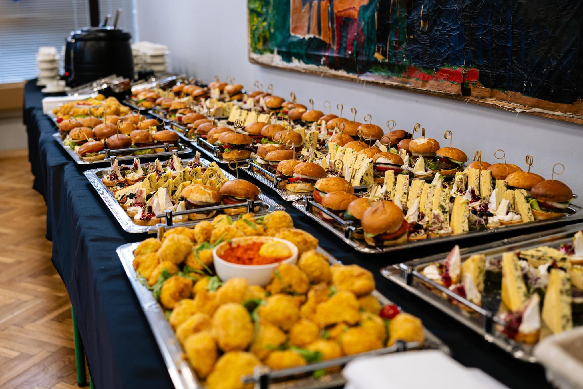 Stół cateringowy z różnorodnymi przekąskami: mini burgery z wykałaczkami, kanapki z serem pleśniowym i bruschetty, wszystko serwowane na srebrnych tacach, z czarnym obrusem i abstrakcyjnym obrazem...