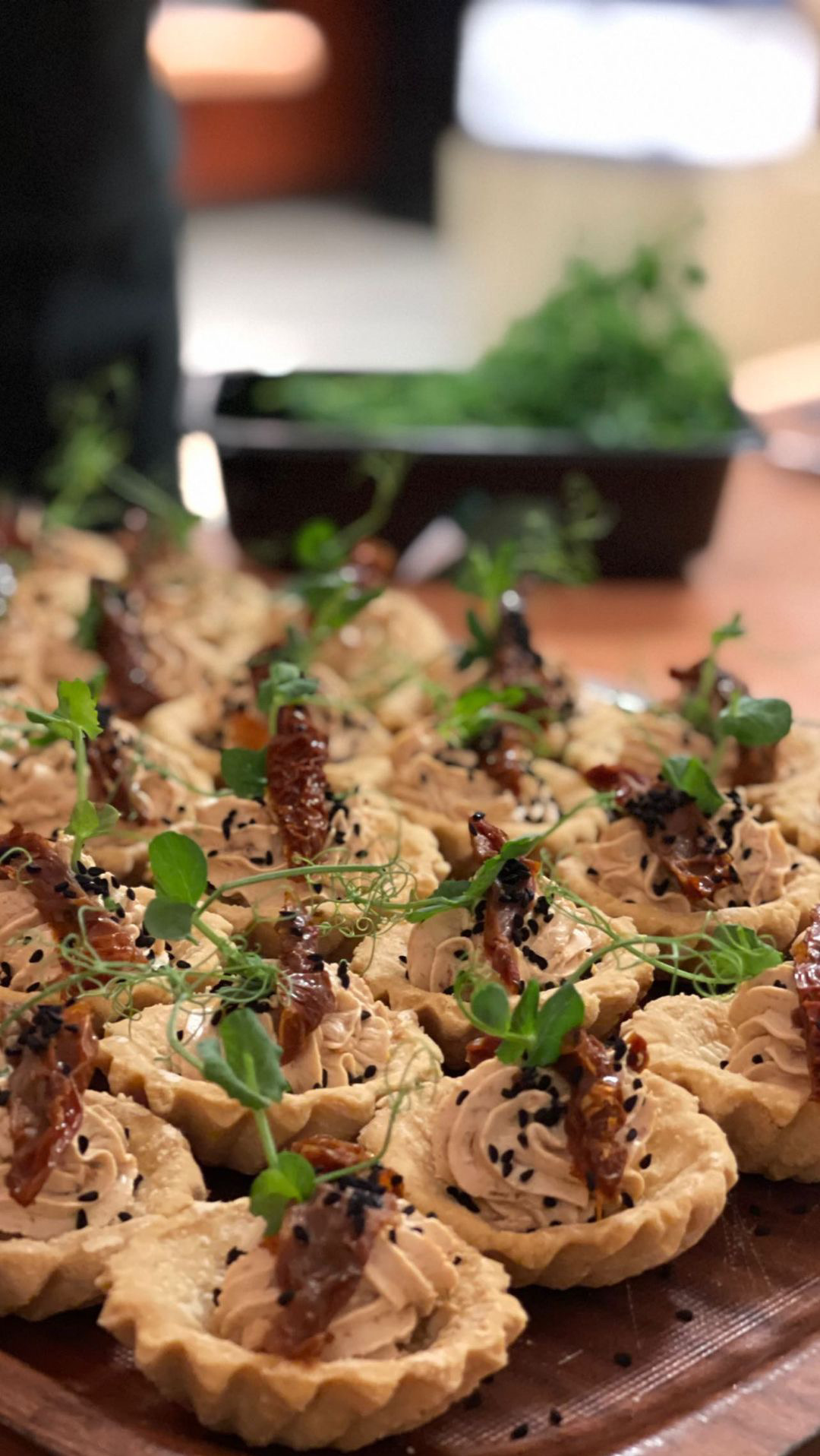 Mini tartaletki z kremowym nadzieniem, suszonymi pomidorami i czarnym sezamem, udekorowane świeżymi listkami, ułożone na drewnianej tacy.