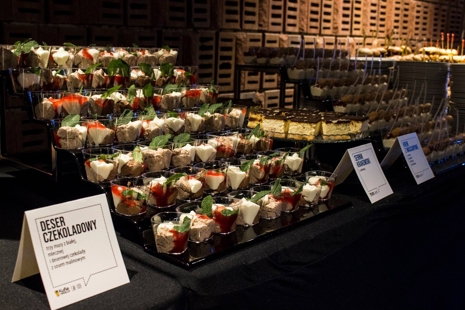 Cateringowy bufet: piramida deserów czekoladowych z sosem malinowym i miętą w plastikowych kubeczkach, obok sernik krakowski i krem z mascarpone, tło z cegieł