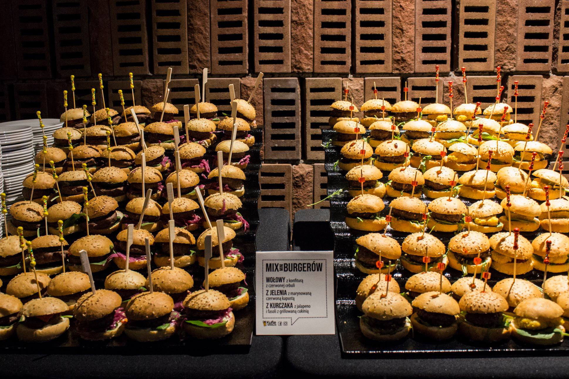 Prezentacja różnorodnych mini burgerów na czarnych tacach, z etykietą 'MIX BURGERÓW' opisującą wołowe, z jelenia i z kurczaka, na tle ściany z cegły.