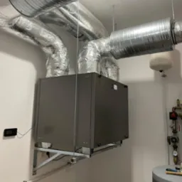 Szara centrala wentylacyjna z podłączonymi przewodami wentylacyjnymi, zamontowana na wspornikach przy ścianie w pomieszczeniu technicznym.