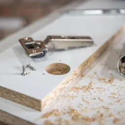 Przygotowanie do montażu zawiasu w białej płycie meblowej, widoczne wiertło, zawias, wkręty i wióry.