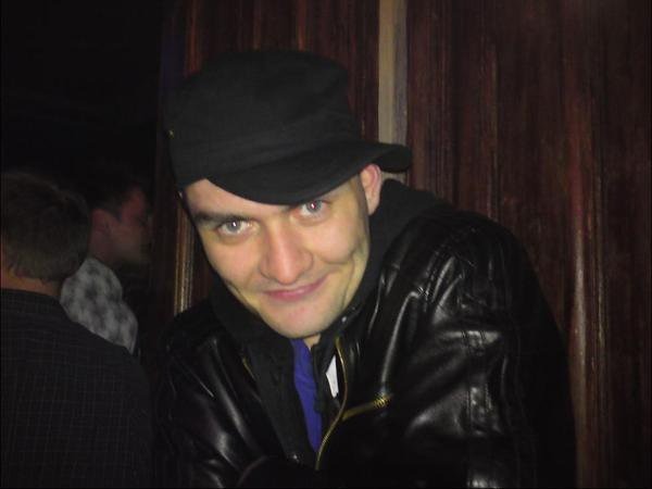 Portret mężczyzny w czarnej skórzanej kurtce i czapce na tle ciemnego, drewnianego panelu.