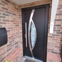 Poldom ltd - Nowoczesne drzwi zewnętrzne w kolorze antracytowym z dekoracyjnym elementem szklanym i srebrną aplikacją, zamontowane w wejściu z ceglaną elewacją.