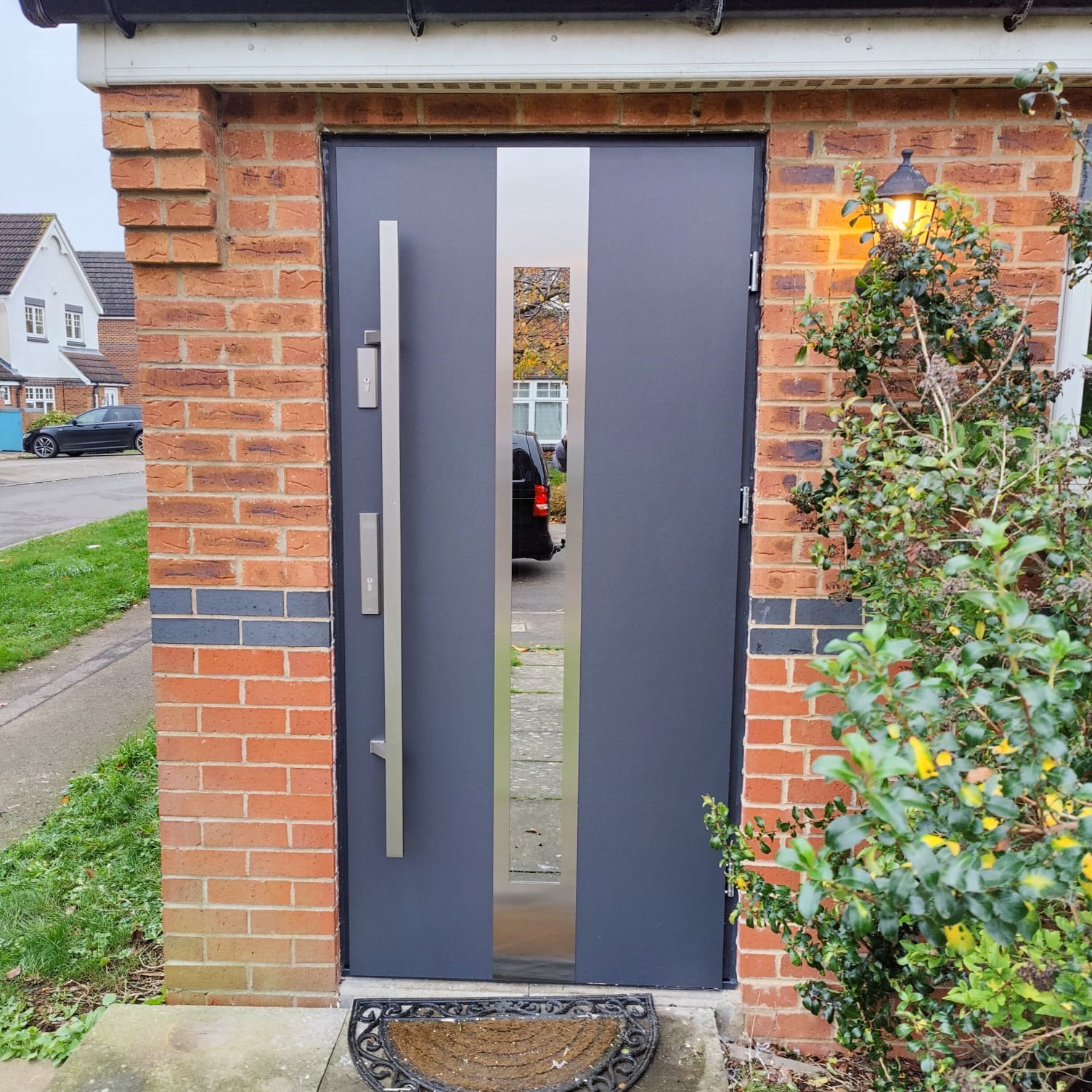 Szare drzwi wejściowe z długim, pionowym uchwytem i lustrzanym panelem centralnym, zamontowane w ceglanej elewacji domu z oświetloną lampą zewnętrzną i wycieraczką przed wejściem.