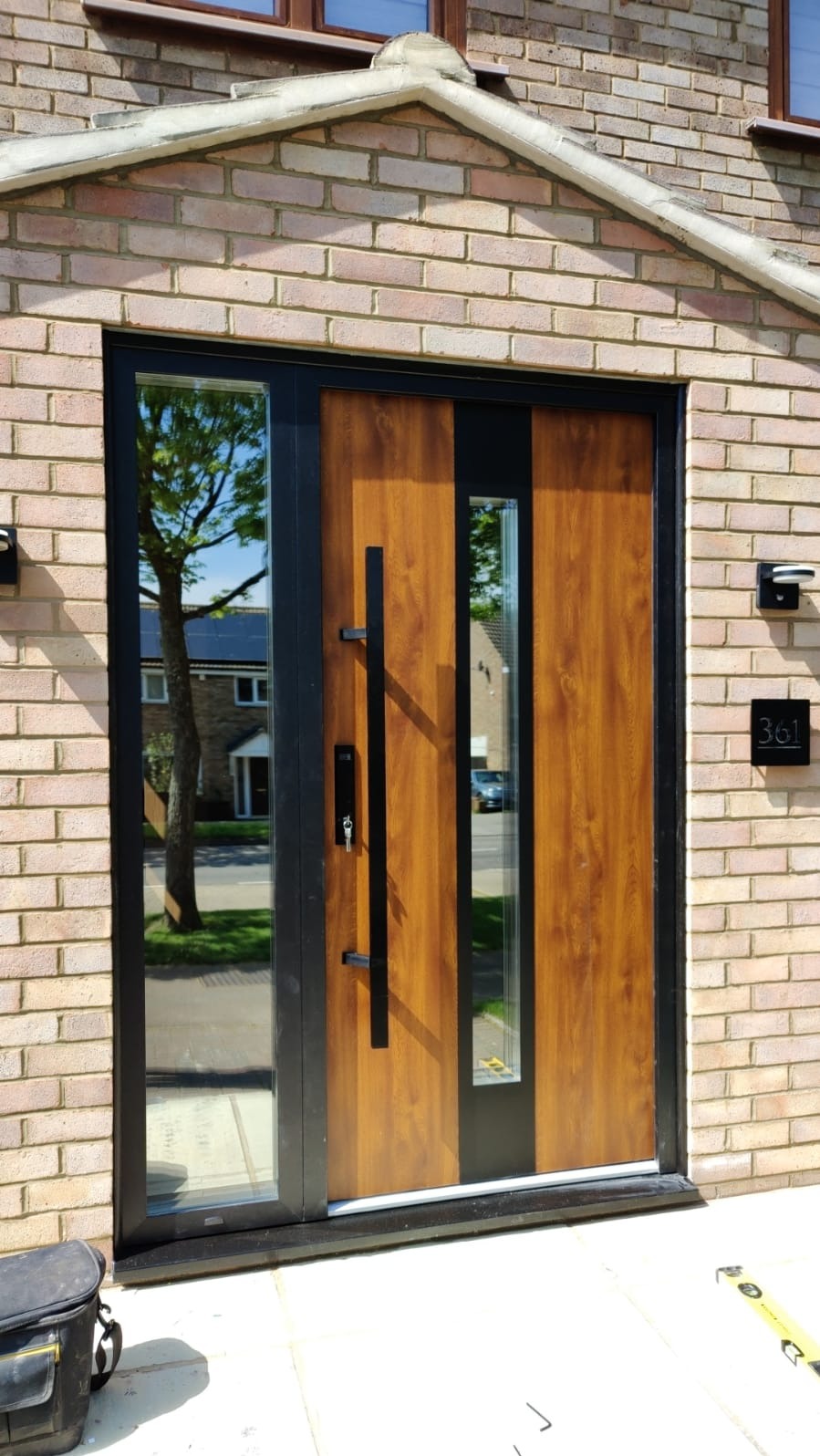 Nowoczesne drzwi wejściowe z panelem drewnopodobnym i szybą, czarna rama, widok z zewnątrz, na pierwszym planie torba narzędziowa i poziomica.