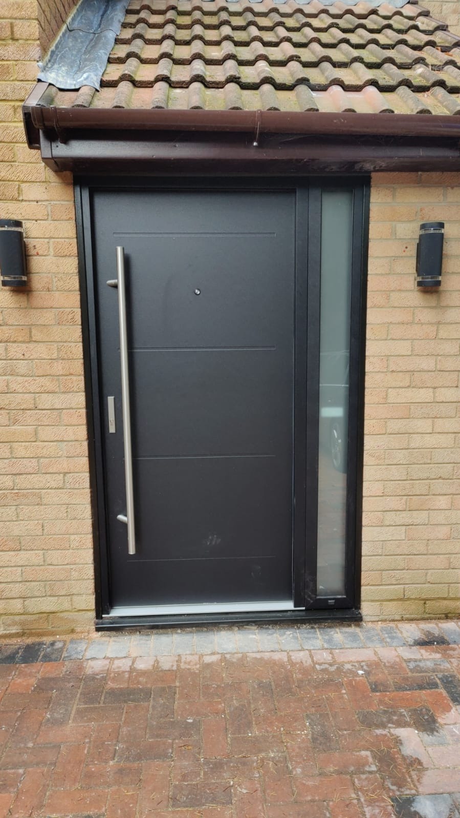 Nowoczesne, czarne drzwi wejściowe z długim, srebrnym uchwytem i bocznym panelem ze szkła matowego, zamontowane w elewacji z cegły klinkierowej pod zadaszeniem z dachówki ceramicznej.