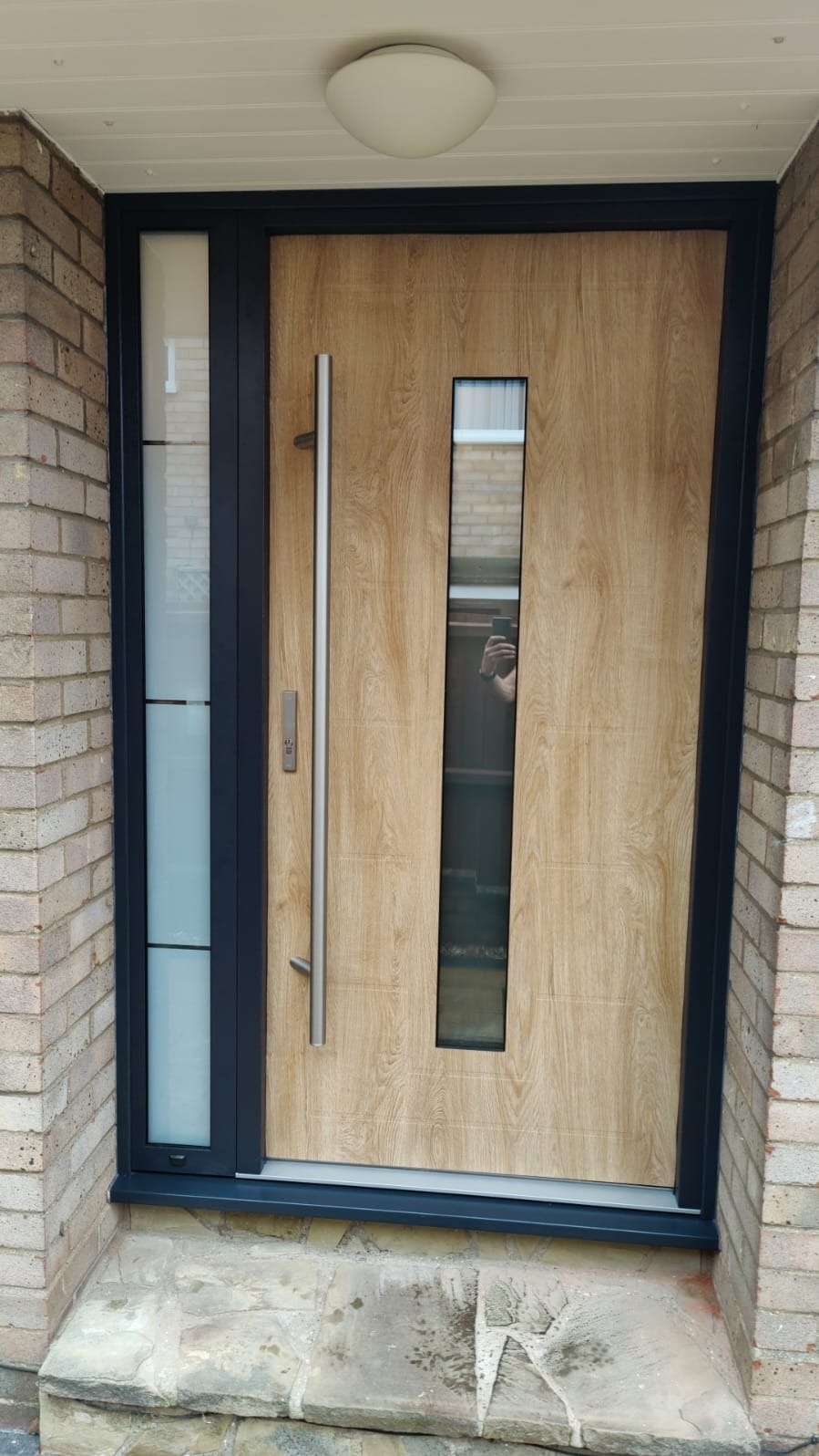 Nowoczesne drzwi wejściowe z drewnopodobnym panelem, długim metalowym uchwytem i pionowym przeszkleniem, osadzone w czarnej ramie, widoczne z zewnątrz na tle ceglanej ściany.