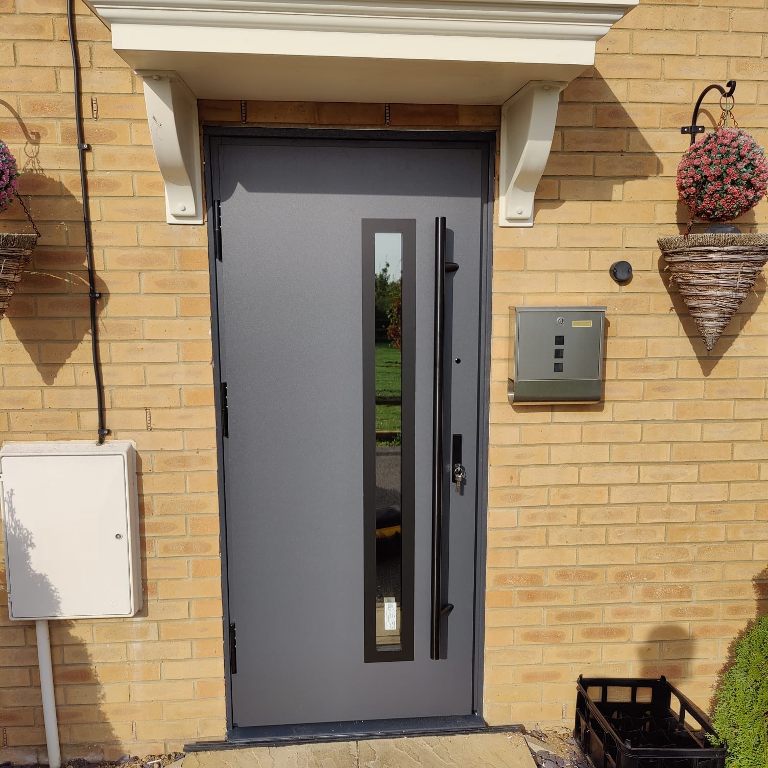 Szare drzwi wejściowe z długim, pionowym przeszkleniem, zamontowane w elewacji z cegły klinkierowej, z dekoracyjnym daszkiem i skrzynką na listy.