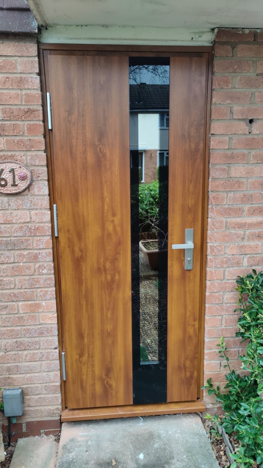 Zewnętrzne drzwi w kolorze drewna z pionowym, czarnym przeszkleniem, zamontowane w ceglanym murze, widoczne z zewnątrz budynku.