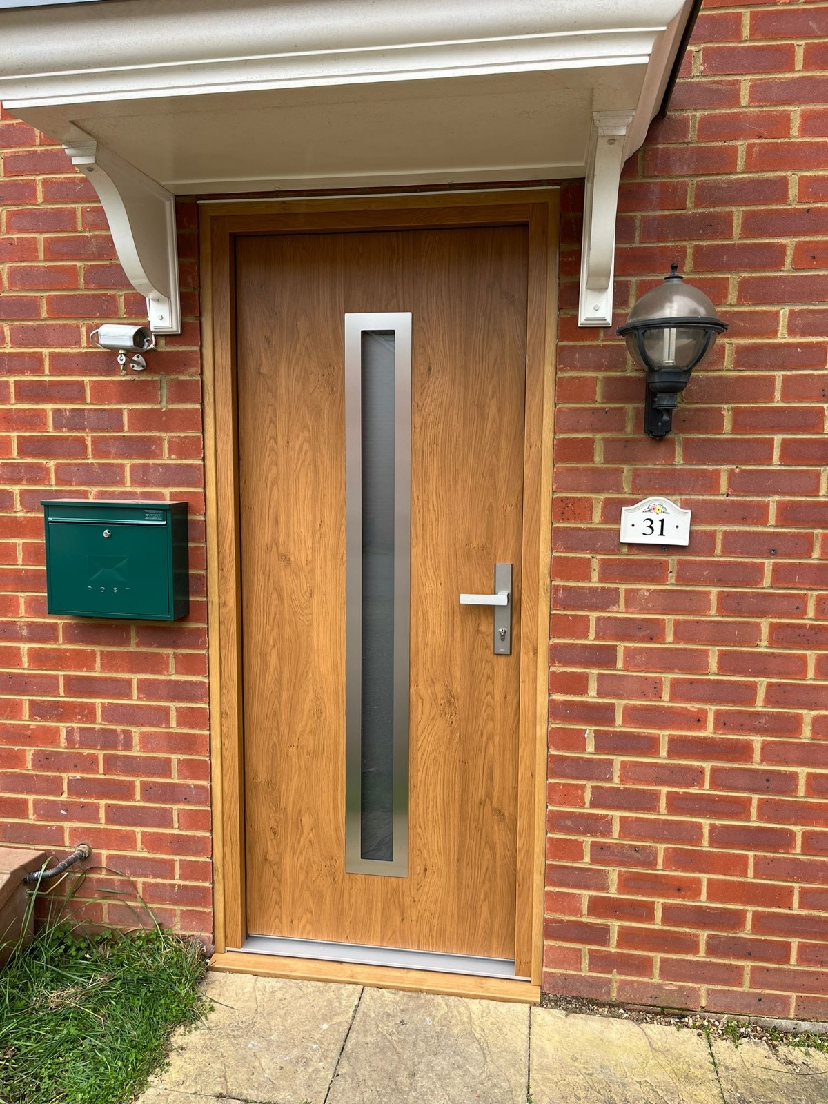 Drewnopodobne drzwi wejściowe z wąskim, pionowym przeszkleniem w metalowej ramie, zamontowane w czerwonej ceglanej ścianie, z zieloną skrzynką na listy i numerem domu '31' obok lampy.