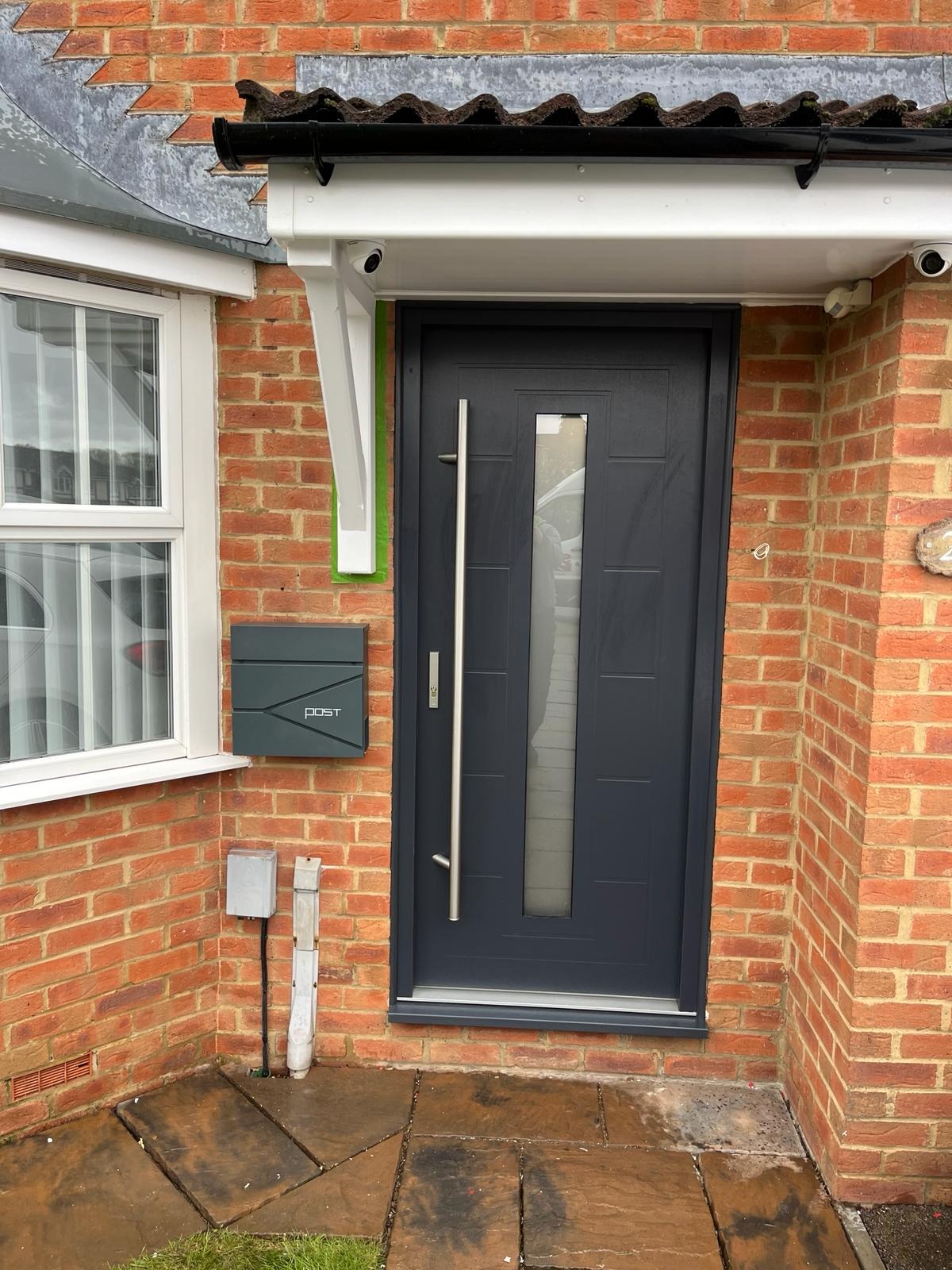 Grafitowe drzwi wejściowe z długim, pionowym uchwytem i szybą, zamontowane w ceglanym budynku z widoczną skrzynką na listy i kamerami monitoringu.