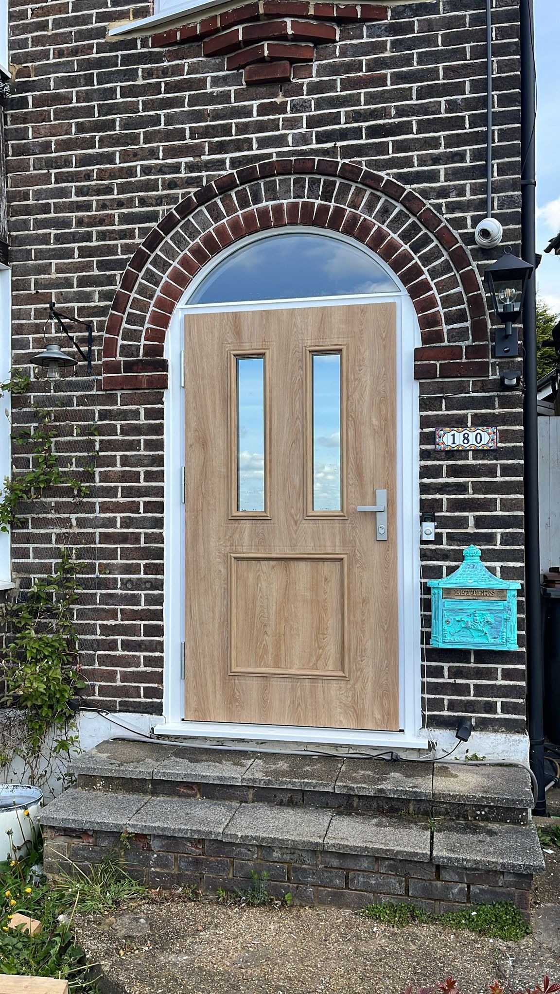 Frontowe drzwi z przeszkleniami w drewnopodobnej okleinie, osadzone w łukowatym portalu z ciemnej cegły, z widoczną skrzynką na listy w stylu vintage i numerem domu 180.
