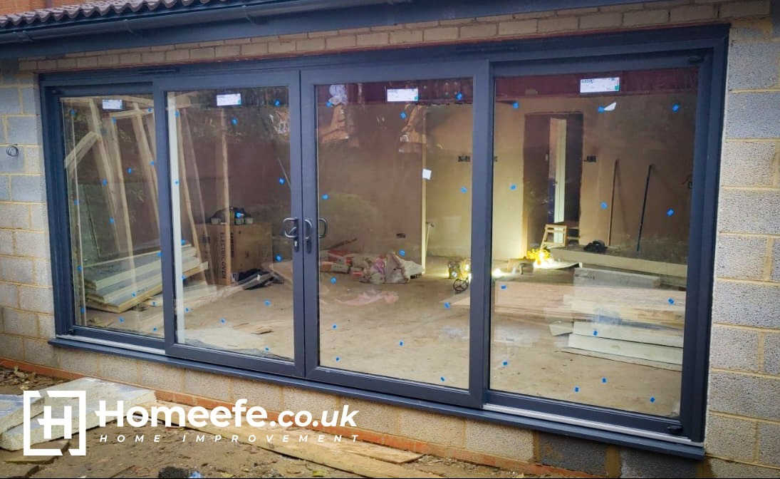 Trzyczęściowe, szare drzwi przesuwne zamontowane w surowym stanie, z widocznymi elementami budowlanymi wewnątrz i niebieskimi zabezpieczeniami na szybach, z logo firmy w dolnej części.