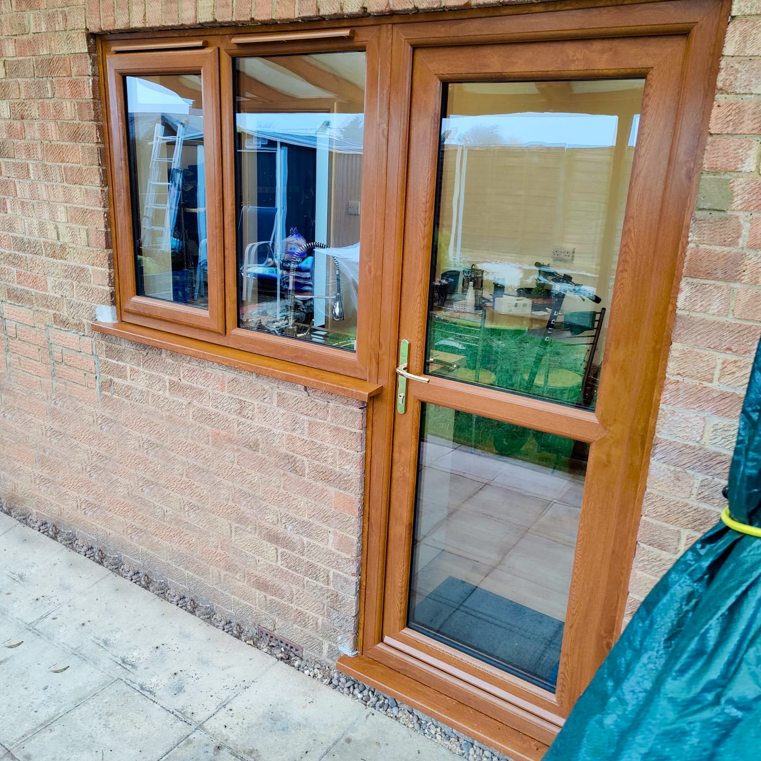 Brązowe okno i drzwi z PCV zamontowane w murze z cegły, widoczne odbicie wnętrza i drabina w oknie, fragment plandeki po prawej stronie.