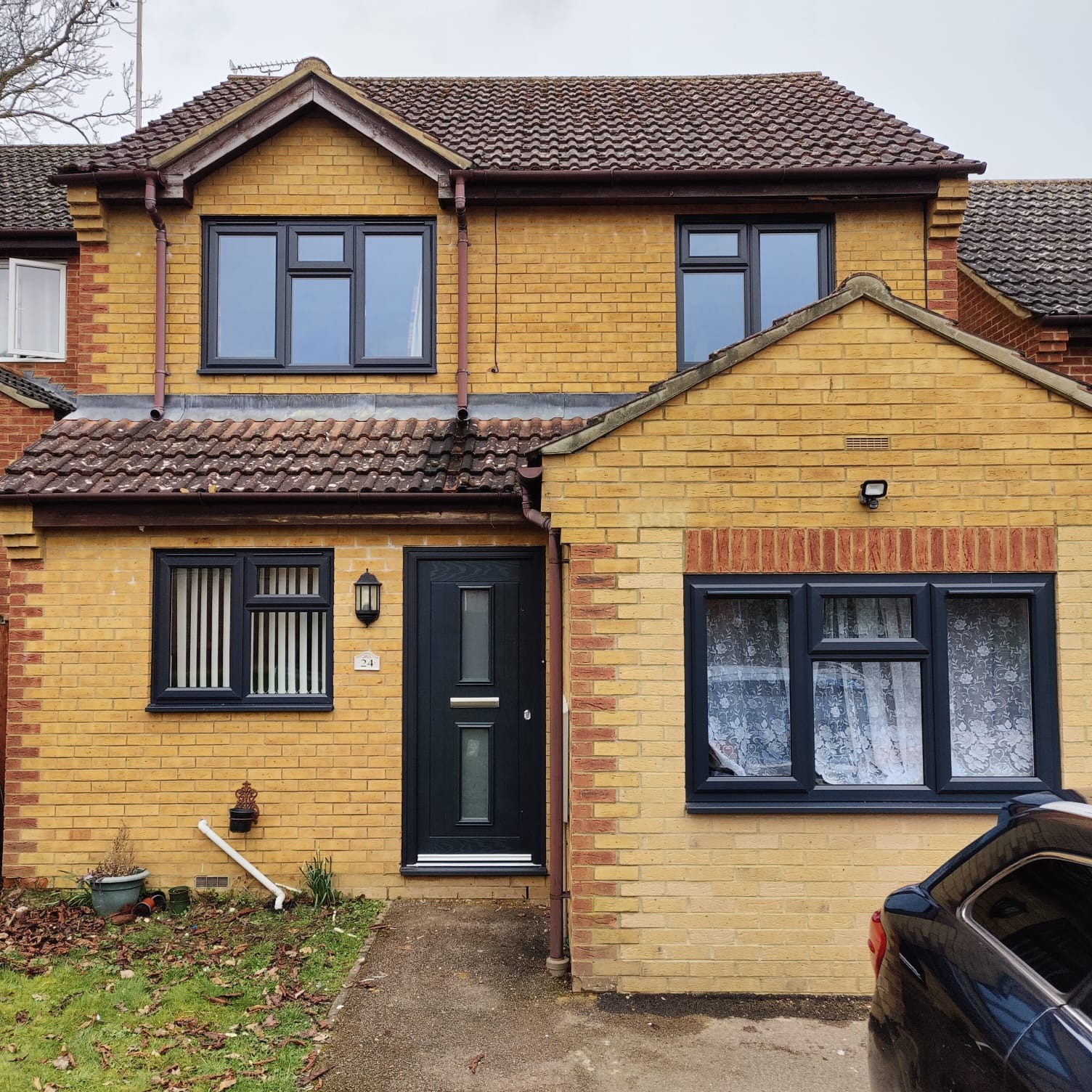 Żółty dom z ciemnoszarą stolarką okienną i drzwiami wejściowymi; widoczne trzy okna na piętrze i dwa na parterze, z czego jedno z żaluzjami pionowymi, a drugie z firanką; drzwi wejściowe z numerem...