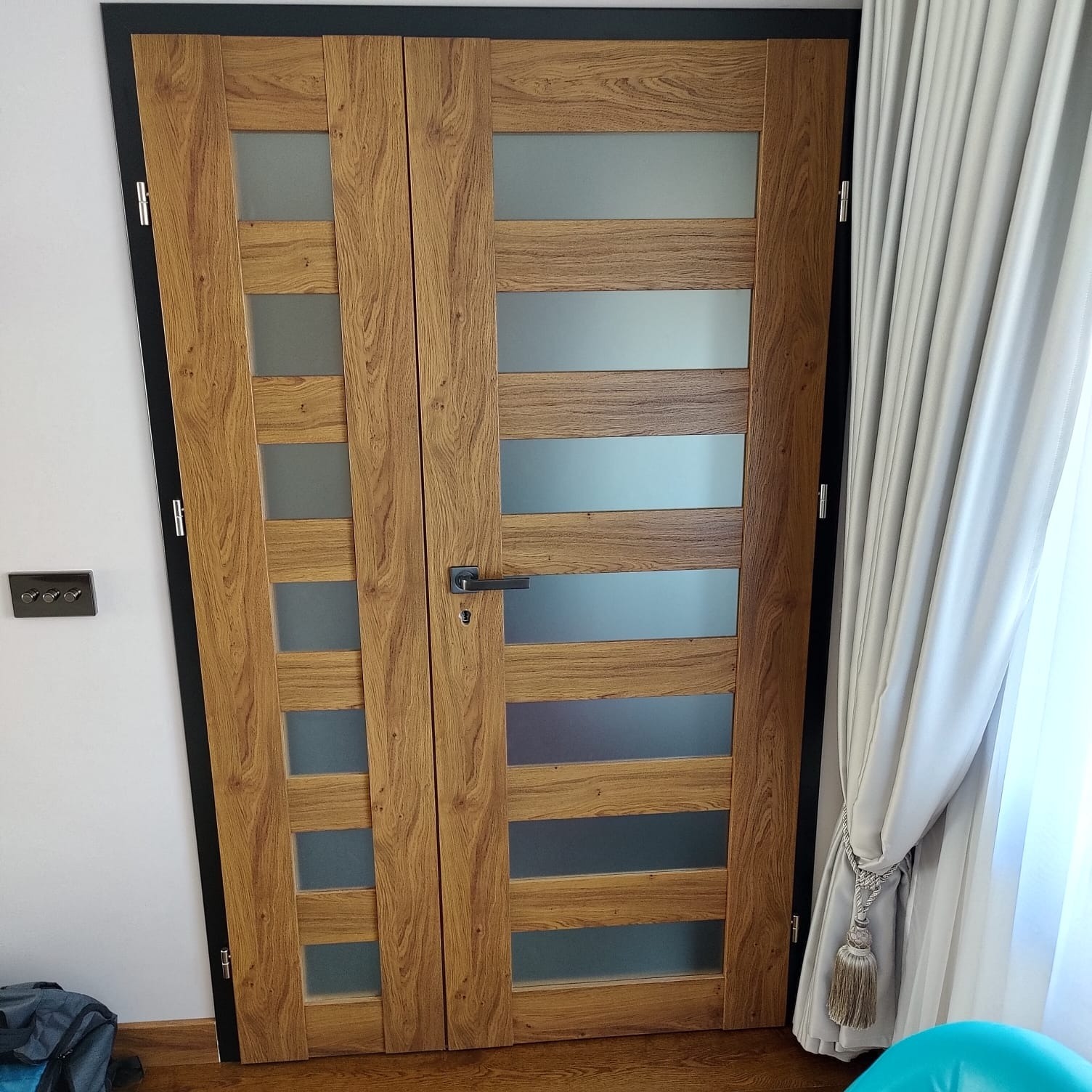 Dwuskrzydłowe drzwi wewnętrzne z drewnianą okleiną i poziomymi, matowymi przeszkleniami, czarna ościeżnica, widoczne firany i włącznik światła.