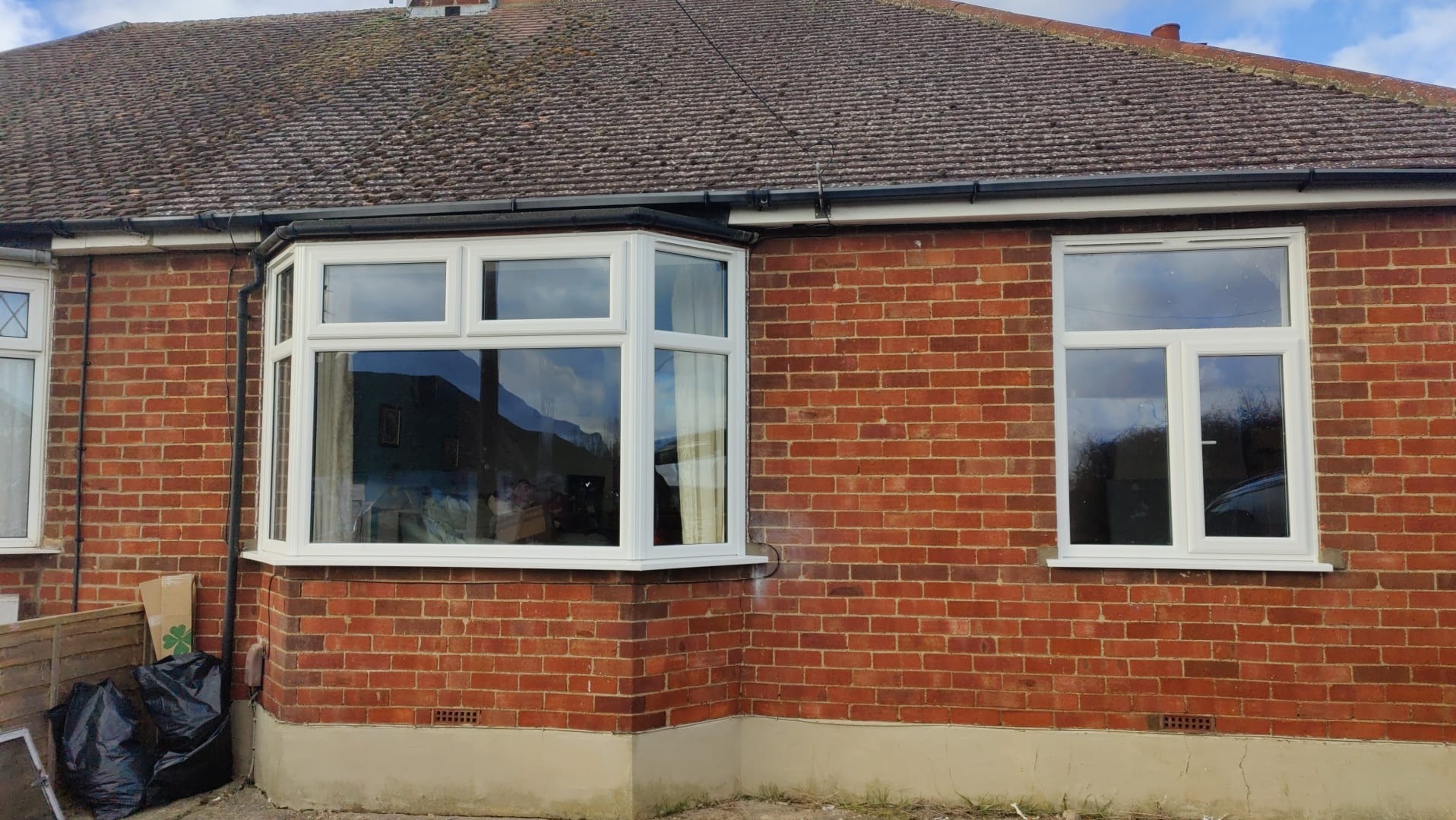 Białe okno wykuszowe i prostokątne okno zamontowane w czerwonej ceglanej ścianie domu, z widocznym fragmentem dachu i niebem odbijającym się w szybach.