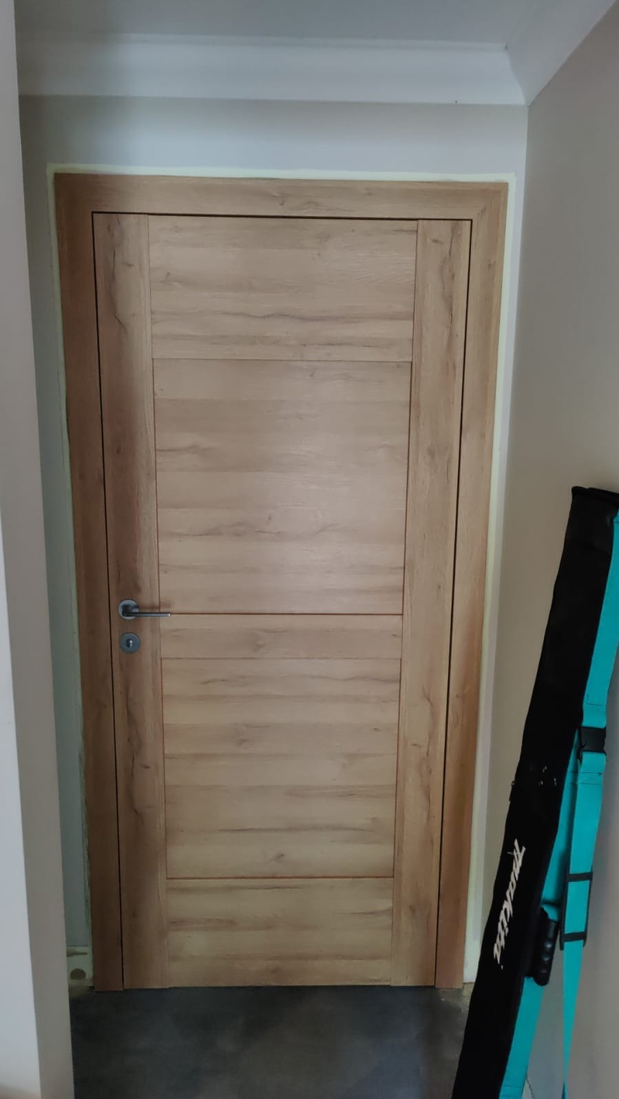 Zamknięte drzwi wewnętrzne z drewnopodobnym wykończeniem i srebrną klamką, obok futerał na narzędzia Makita.