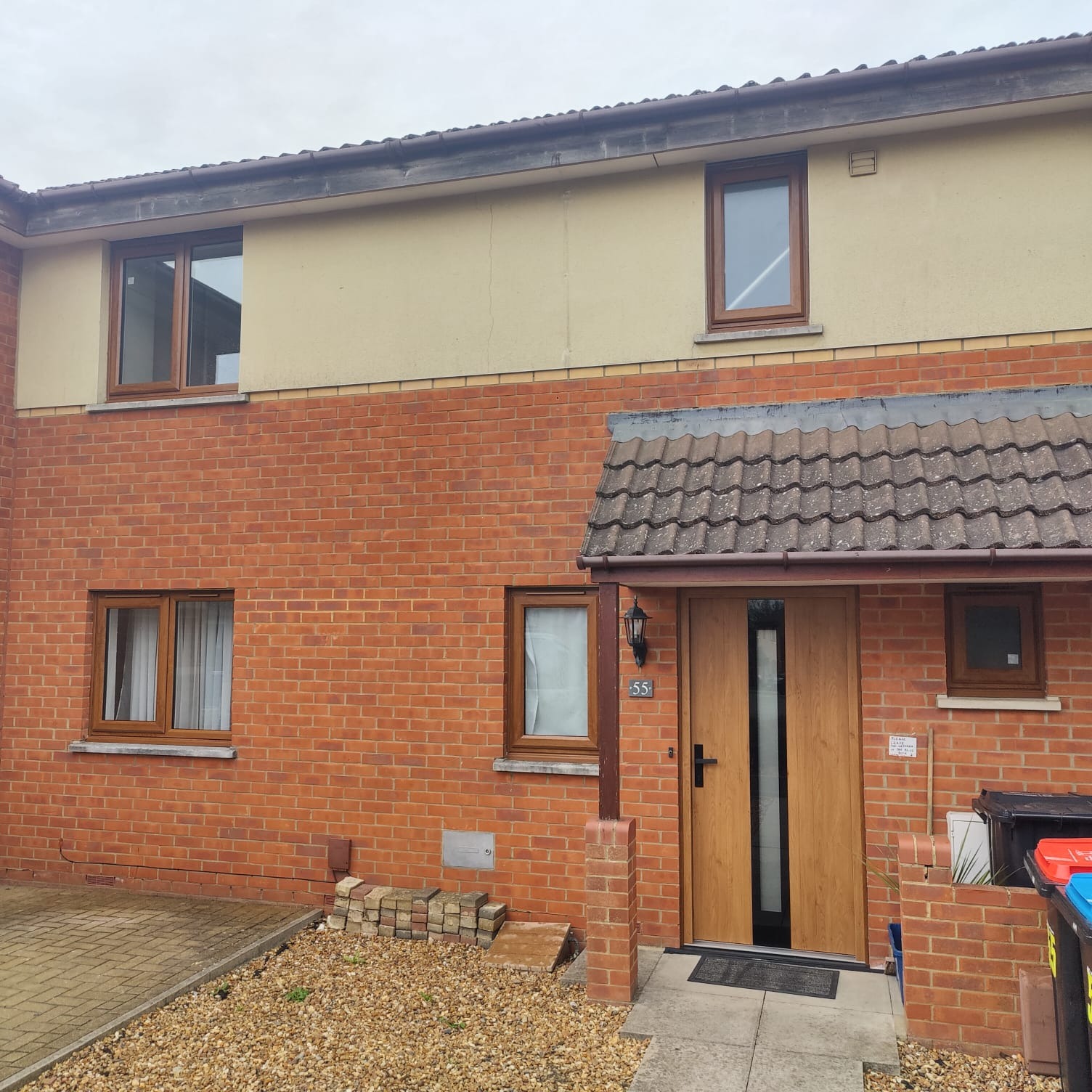 Fasada budynku z cegły z brązowymi oknami i drzwiami wejściowymi z numerem 55, widoczne kosze na śmieci i stos cegieł.