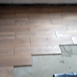 Złota rączka - Układanie paneli podłogowych w trakcie pracy, widoczne listwy dystansowe i folia ochronna na podłodze, surowy stan wykończenia ścian i sufitu.