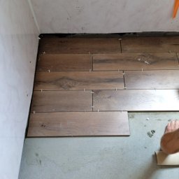 Złota rączka - Układanie drewnopodobnych płytek podłogowych z użyciem krzyżyków dystansowych; widoczny fragment stopy i rolka taśmy malarskiej.