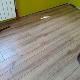 Złota rączka - Ułożona podłoga z paneli laminowanych w jasnym odcieniu drewna, widoczny grzejnik i żółta ściana w tle.